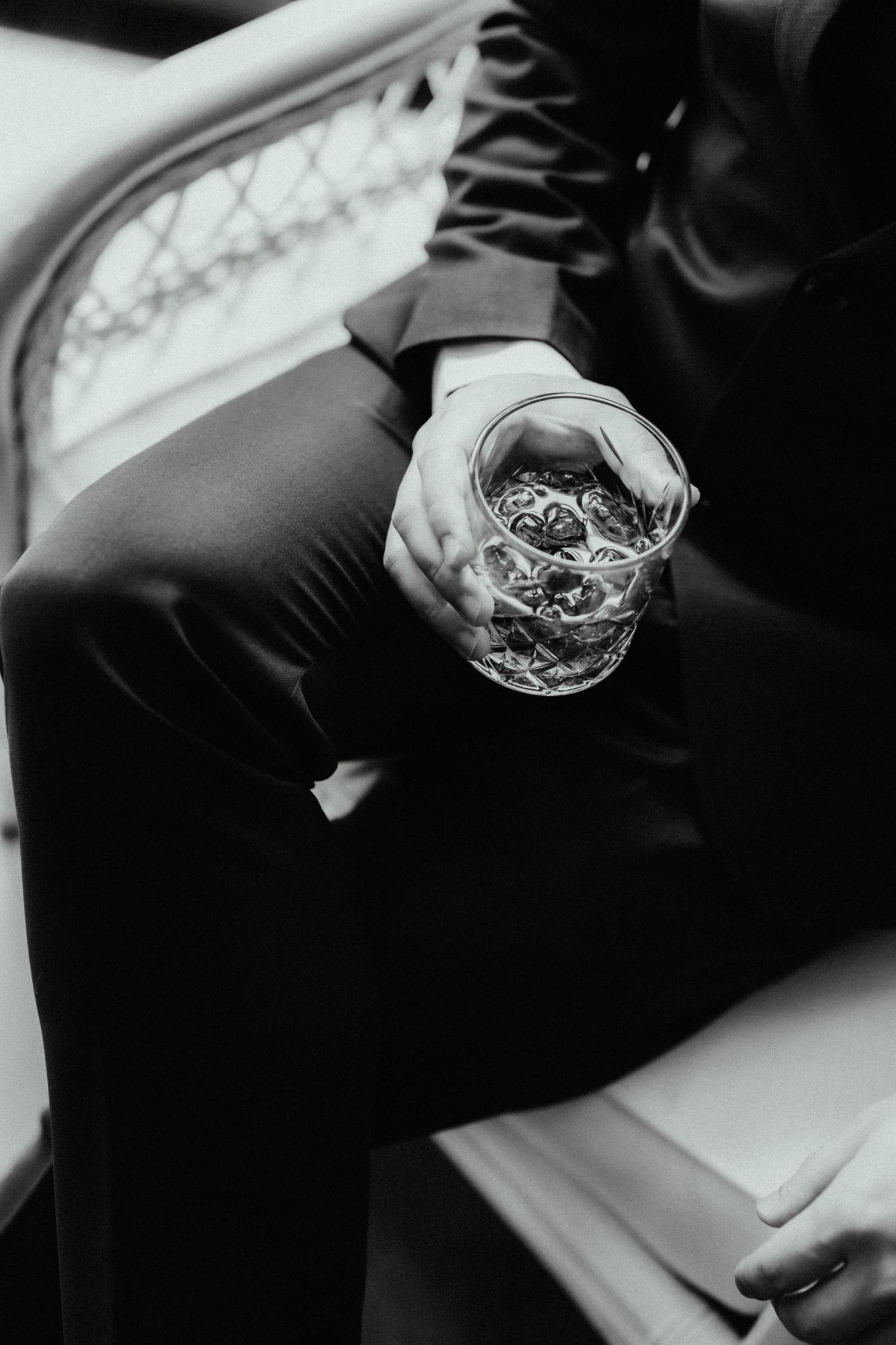 Black and white photo of a man in a dark suit seated on a wicker chair, holding a crystal glass of whiskey in his hand.