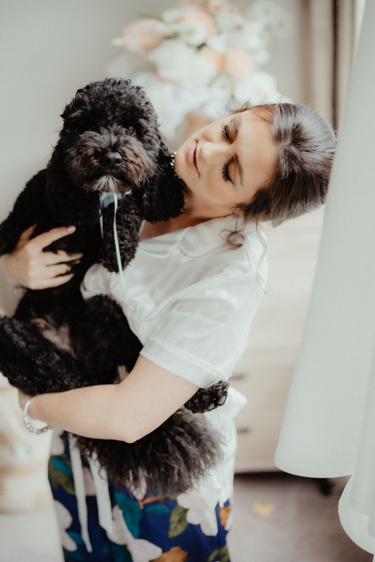 Woman in a white shirt smiles while holding a fluffy black dog with a bow tie, with a floral bouquet in the soft-lit background.