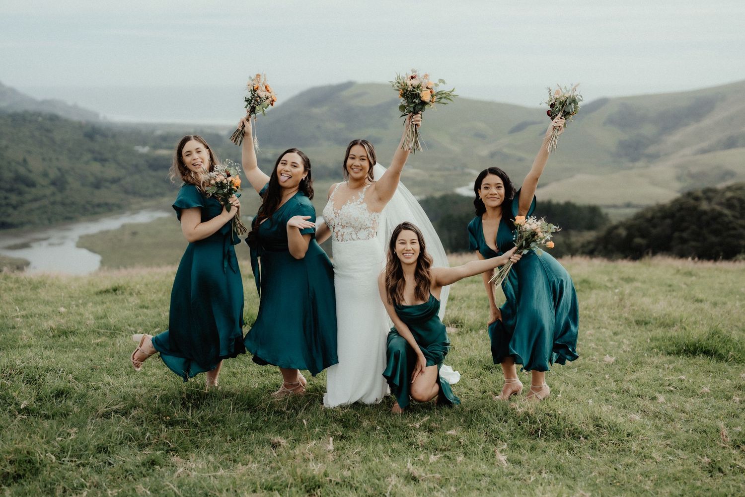 Bride and four bridesmaids in teal dresses lift their bouquets and laugh together on a grassy hill with rolling views behind.