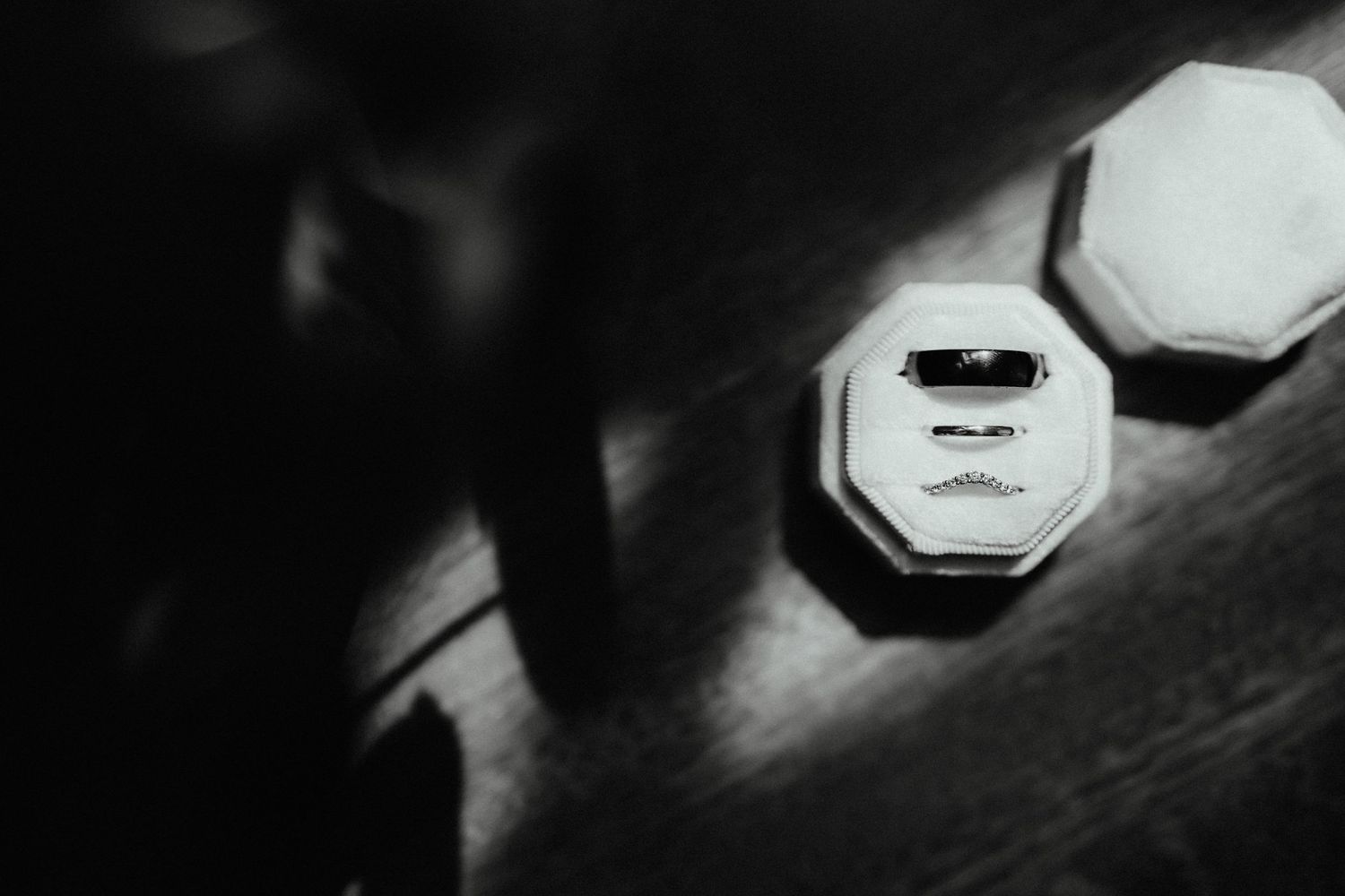Black and white photo of an open octagonal ring box holding wedding bands on a wooden table beside its matching lid.