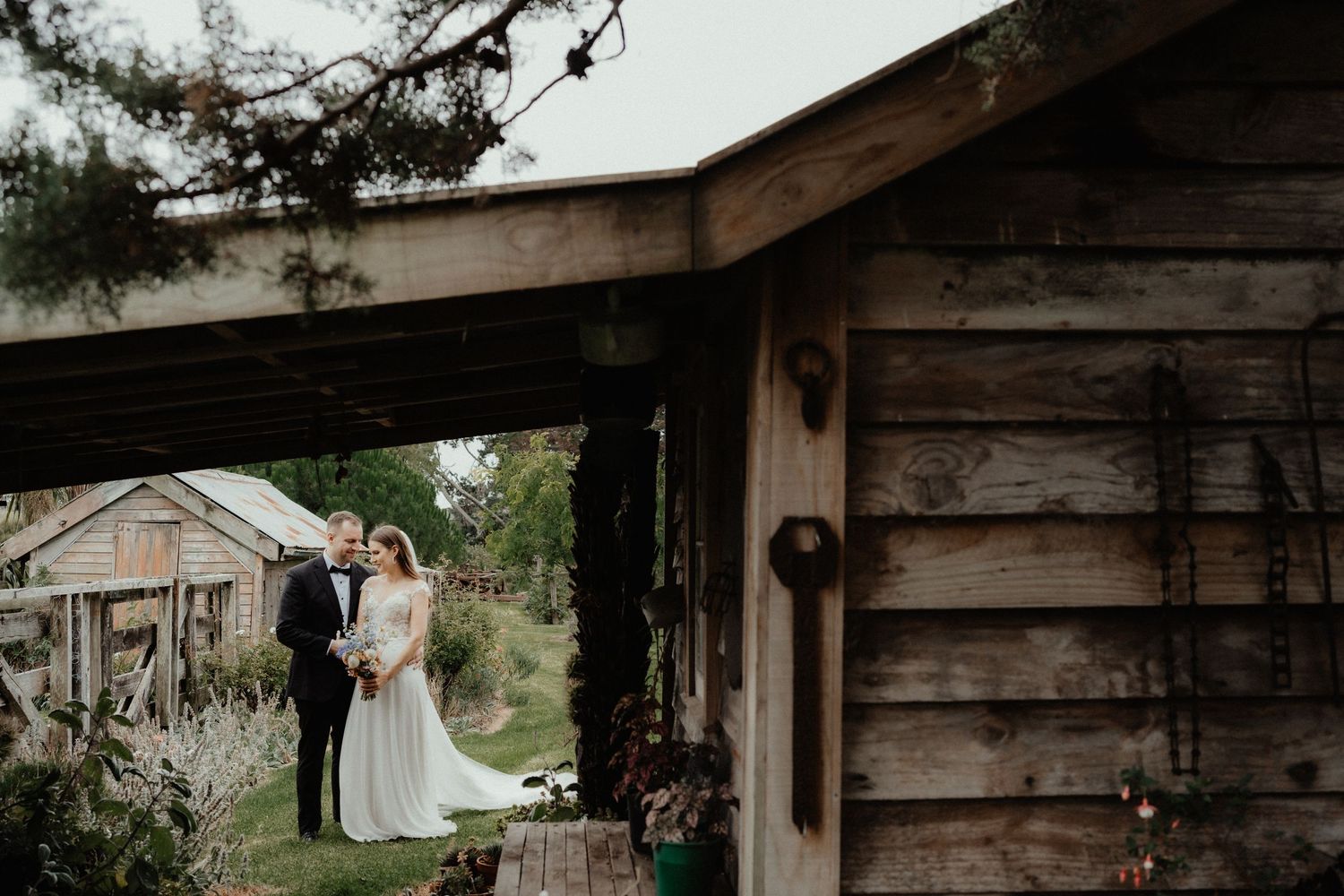 Newlyweds pose side by side beside a weathered wooden building with a lush garden and old farm sheds nearby.