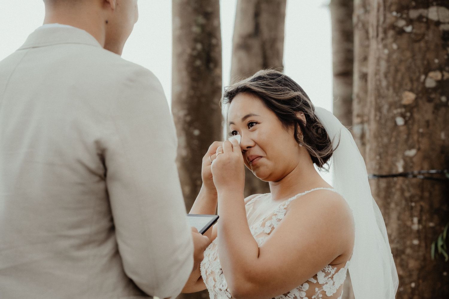 Bride in a lace gown and veil wipes away happy tears while exchanging vows with her groom among tall trees.