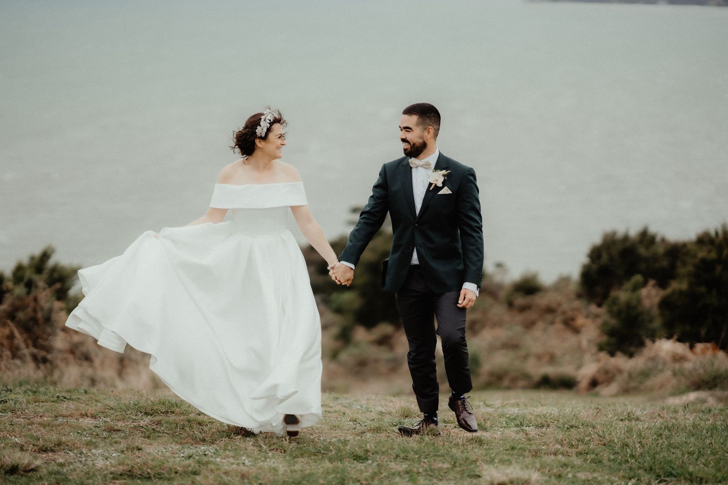 Bride and groom hold hands and walk across a grassy clifftop above the sea, her white off-shoulder gown flowing in the breeze.
