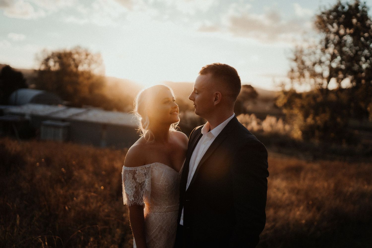 Bride and groom crouch to greet a spotted dog while smiling wedding guests gather around them in a forest clearing.