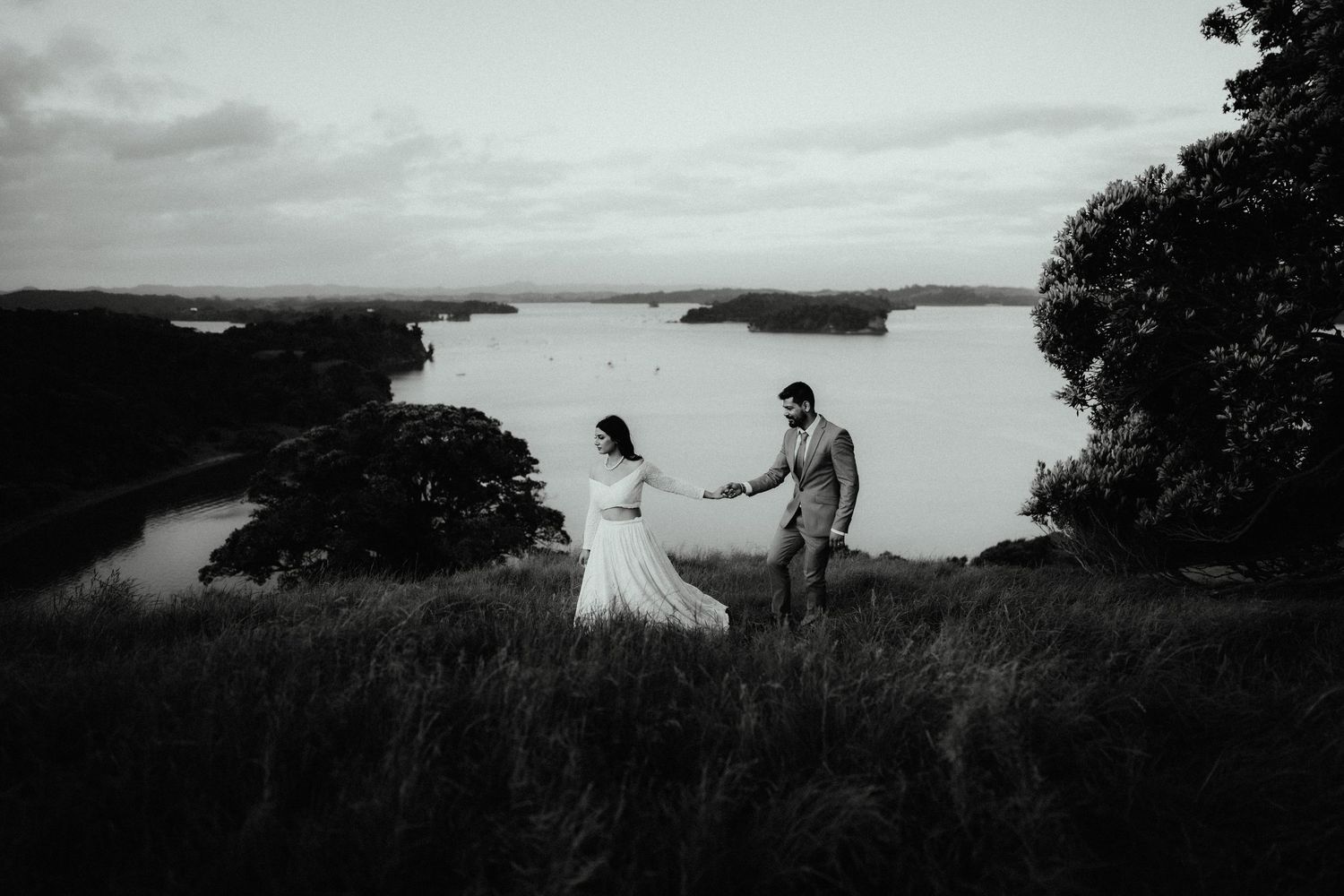 Black and white wide shot of a bride and groom holding hands on a grassy hillside above a scenic coastal bay.