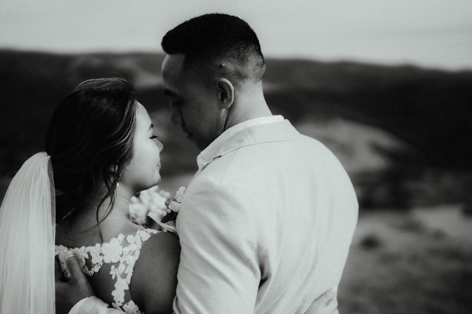 Black and white close-up of a bride in a lace gown and veil gazing at her groom outdoors on their wedding day.