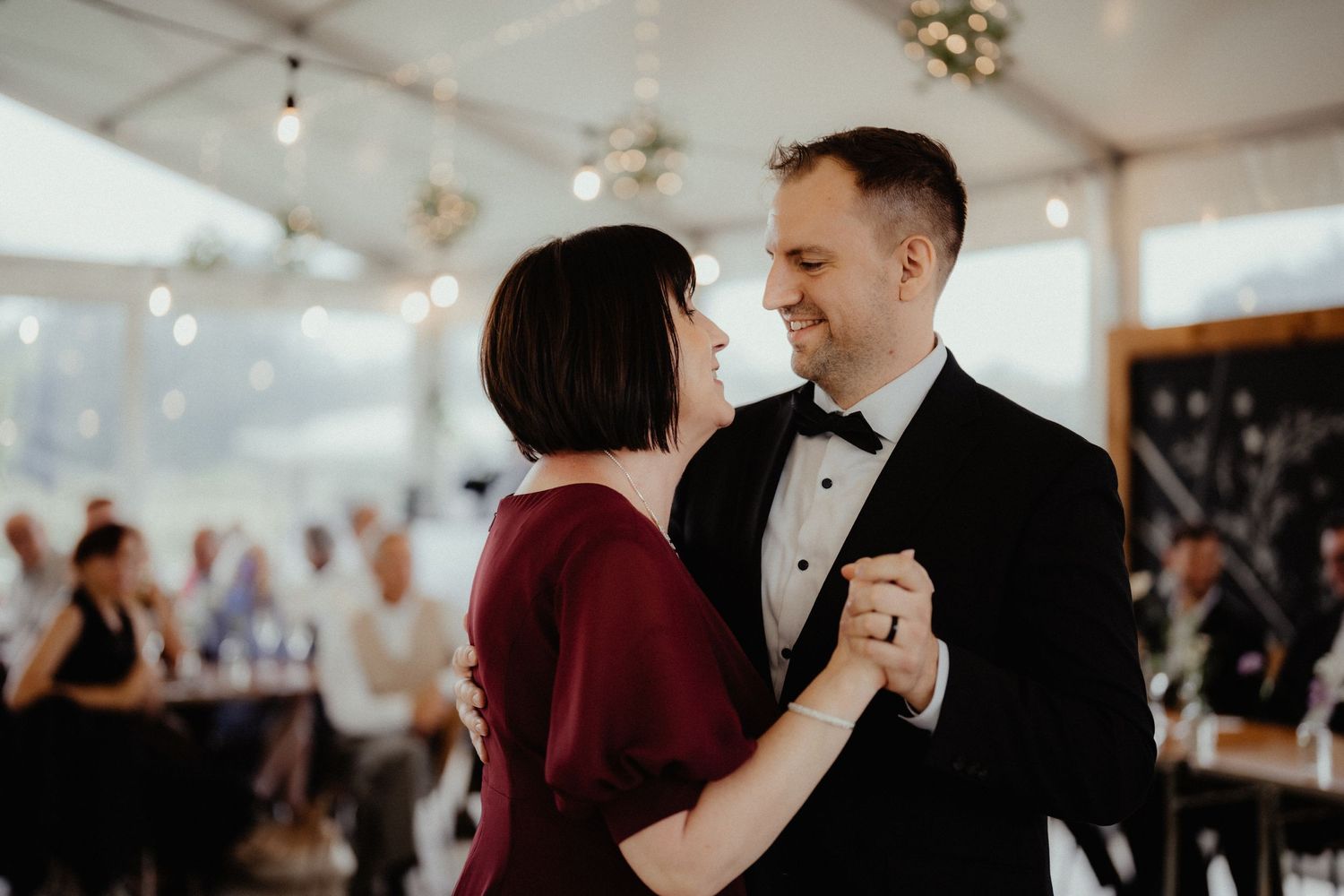 Smiling groom in a black tuxedo and bow tie dances with a woman in a burgundy dress while wedding guests look on in a marquee.