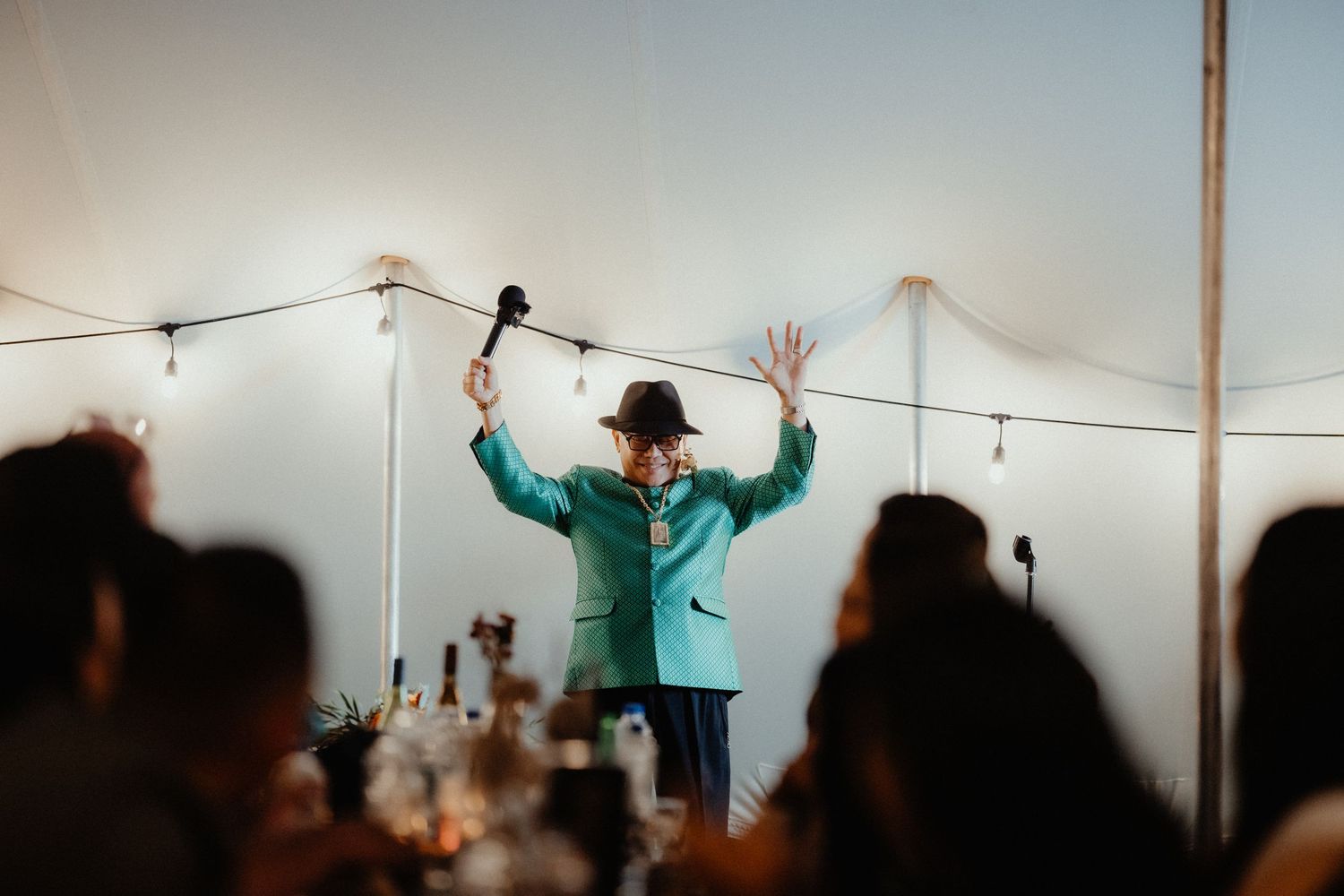 A man in a teal jacket and dark hat raises his arms and holds a microphone while giving an animated wedding reception speech.
