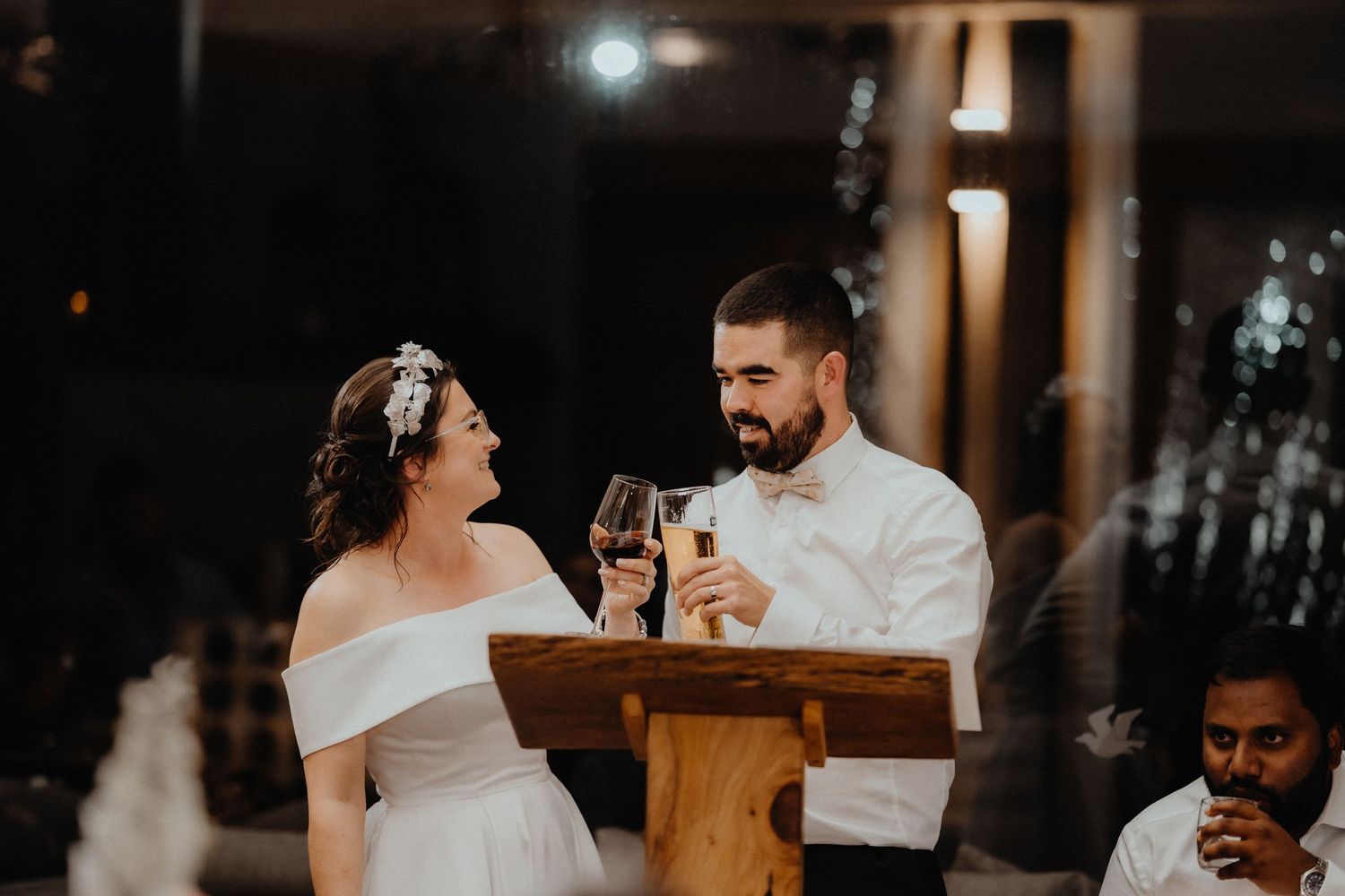 Bride and groom share a champagne toast at a wooden lectern during their wedding reception speeches.