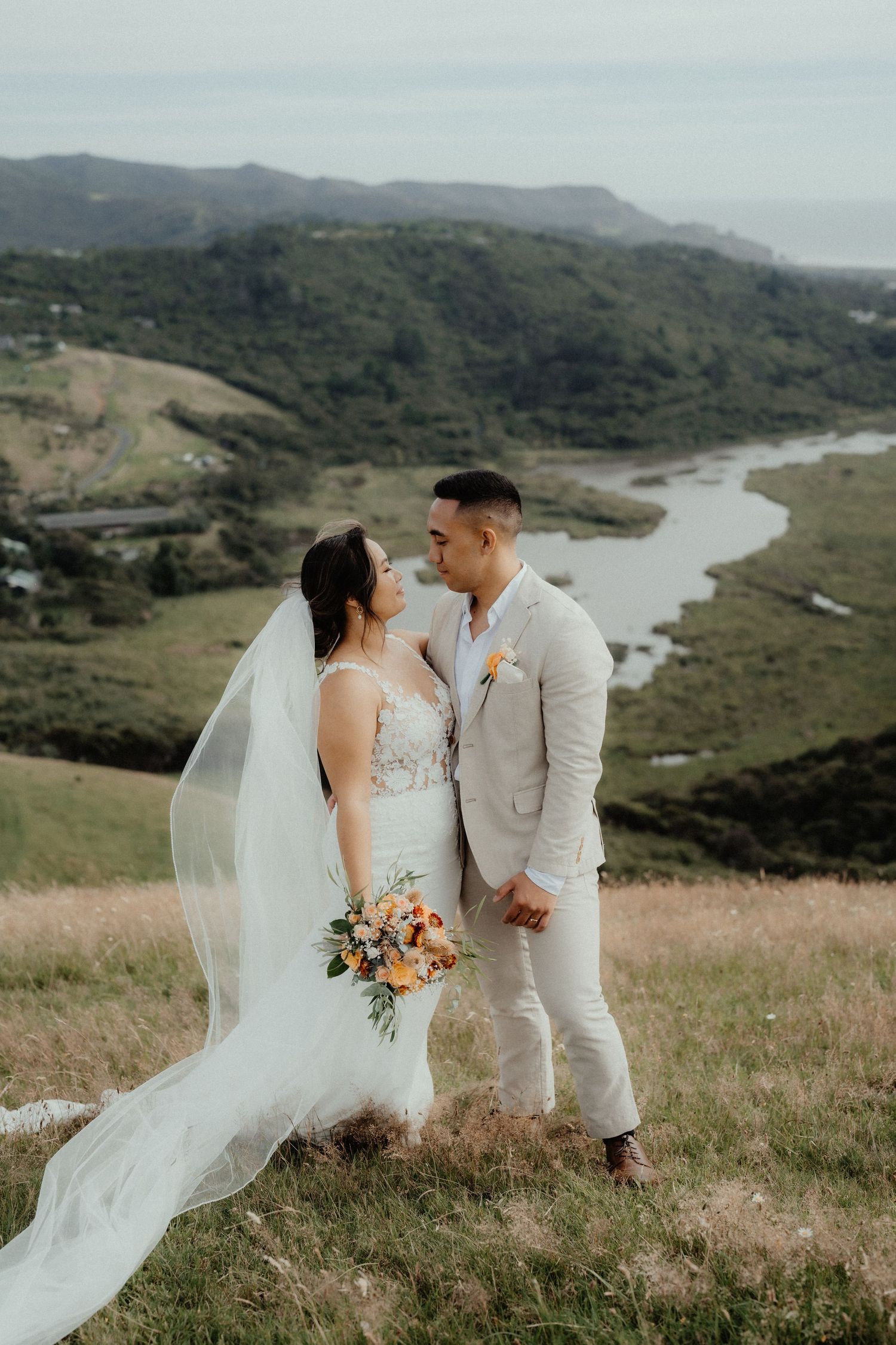 Bride and groom share an intimate moment on a hilltop wedding day with a bouquet, flowing veil and a scenic river valley behind them.