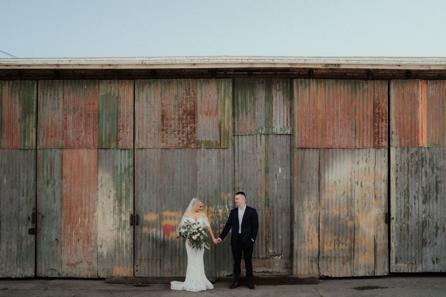 Bride in a white gown holding a bouquet stands beside a groom in a navy suit in front of a rustic corrugated iron barn.