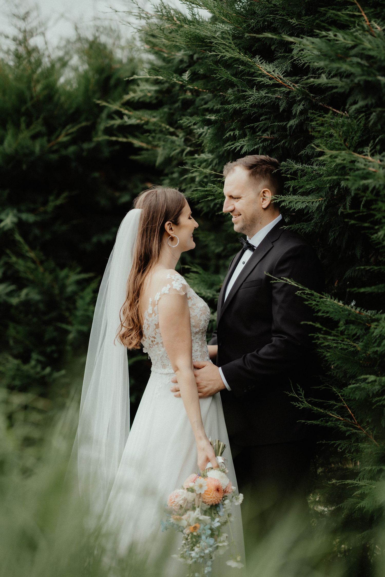 Bride in a lace gown and long veil gazes lovingly at her groom while holding a pastel bouquet among evergreen trees.