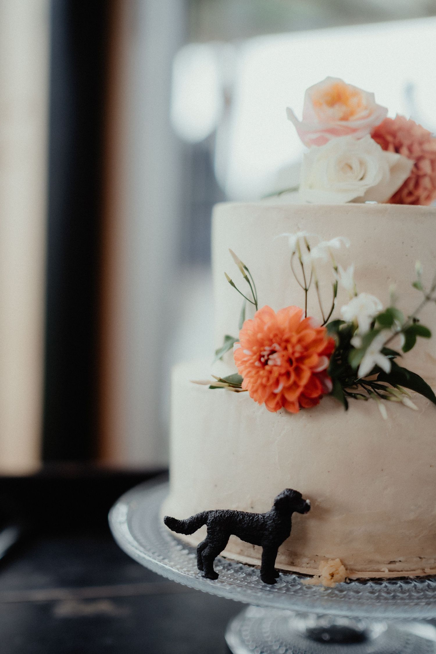 Two-tier white wedding cake decorated with peach roses, an orange dahlia and greenery, topped with a small black dog figurine.
