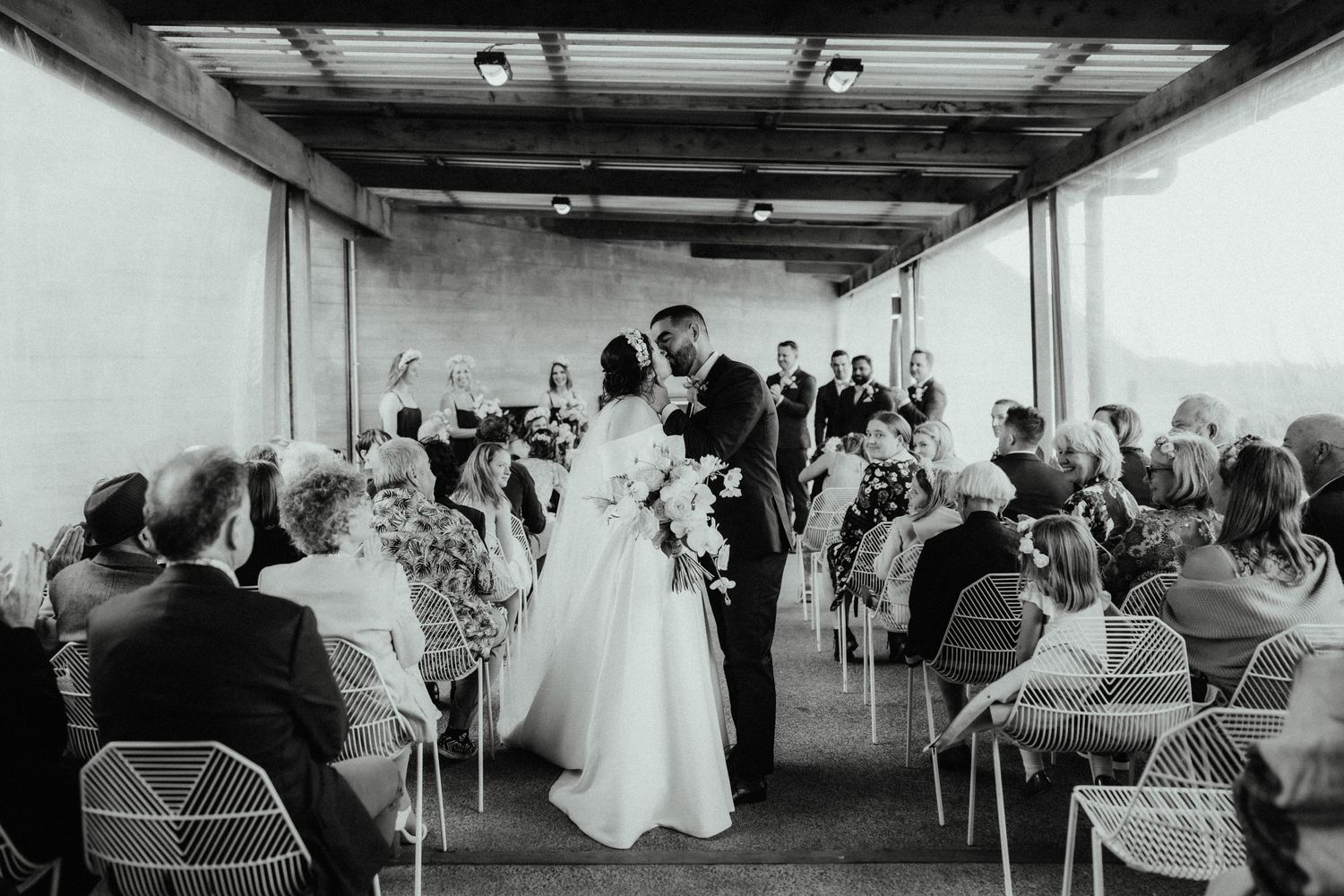 A bride and groom share a kiss in the aisle surrounded by seated wedding guests in a black and white photo.