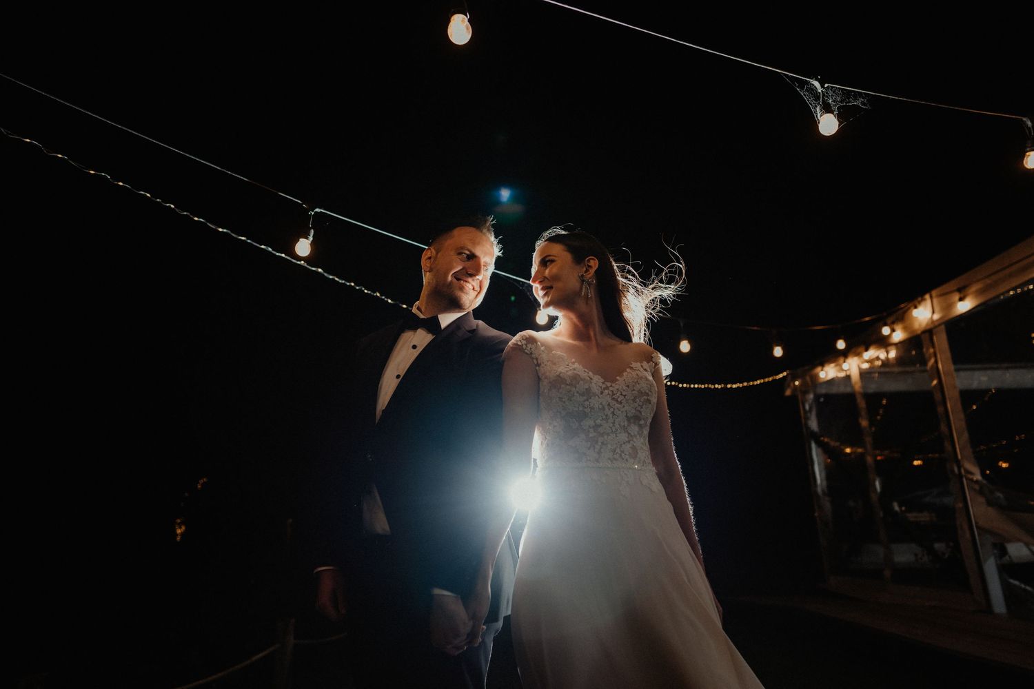Bride and groom smile at each other at night under string lights, the bride in a lace wedding gown with a glowing light between them.