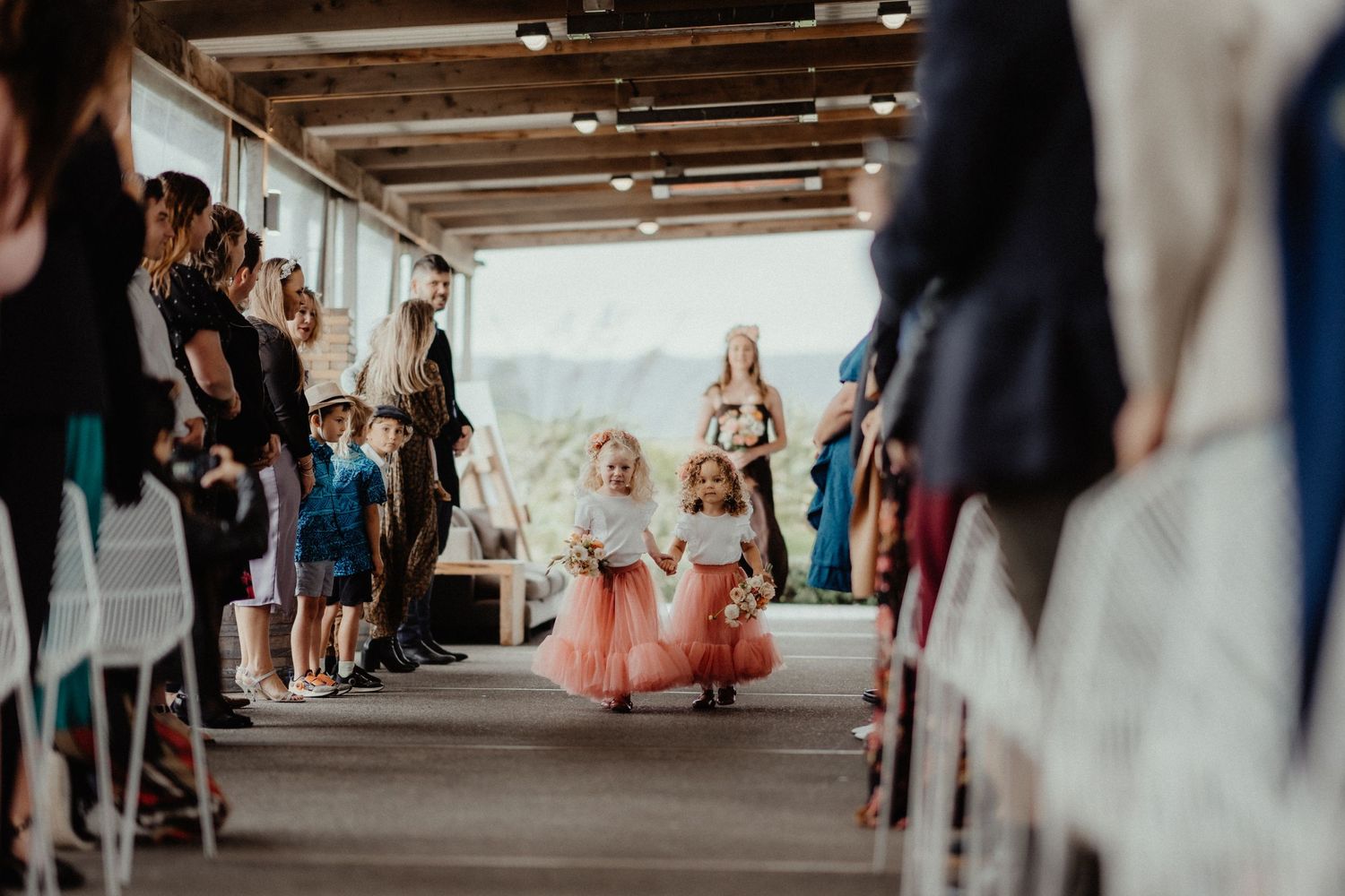 Two little flower girls in pink tulle skirts walk down the aisle between rows of seated guests at a wedding ceremony.