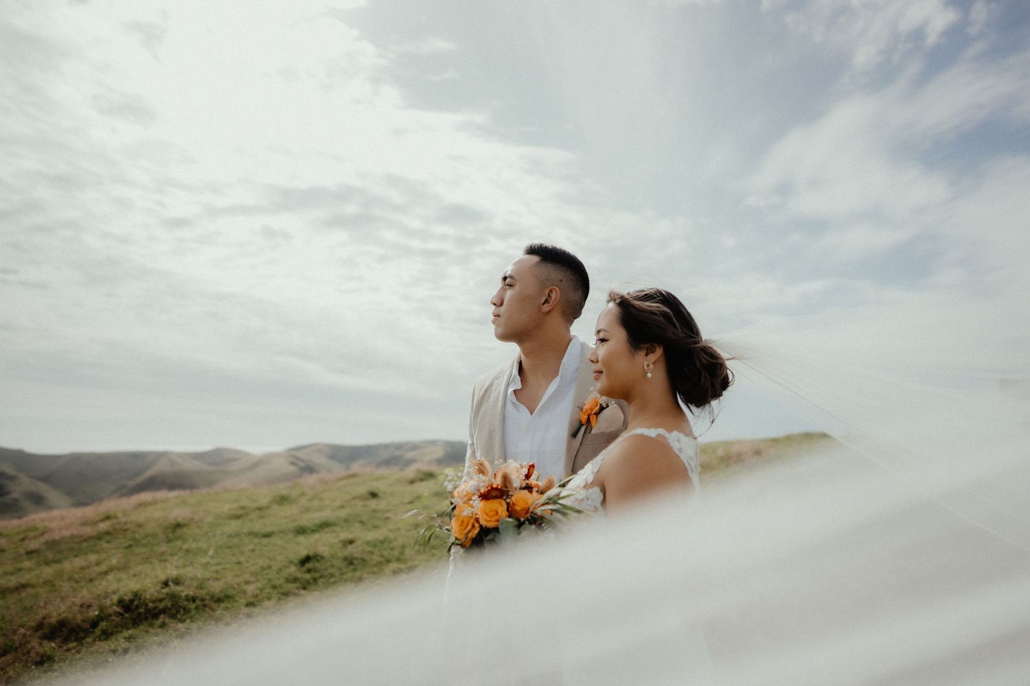 A bride and groom hold an orange bouquet and look out over green hills under a cloudy sky, seen through a flowing veil.