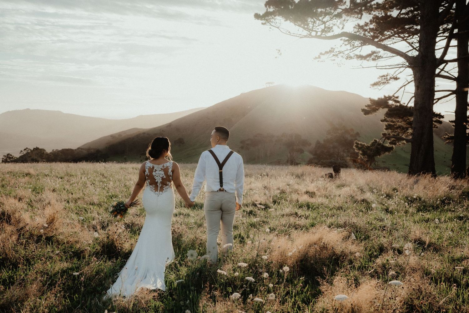 Bride and groom walk hand in hand through golden grass toward rolling hills as the sun sets at their countryside wedding.
