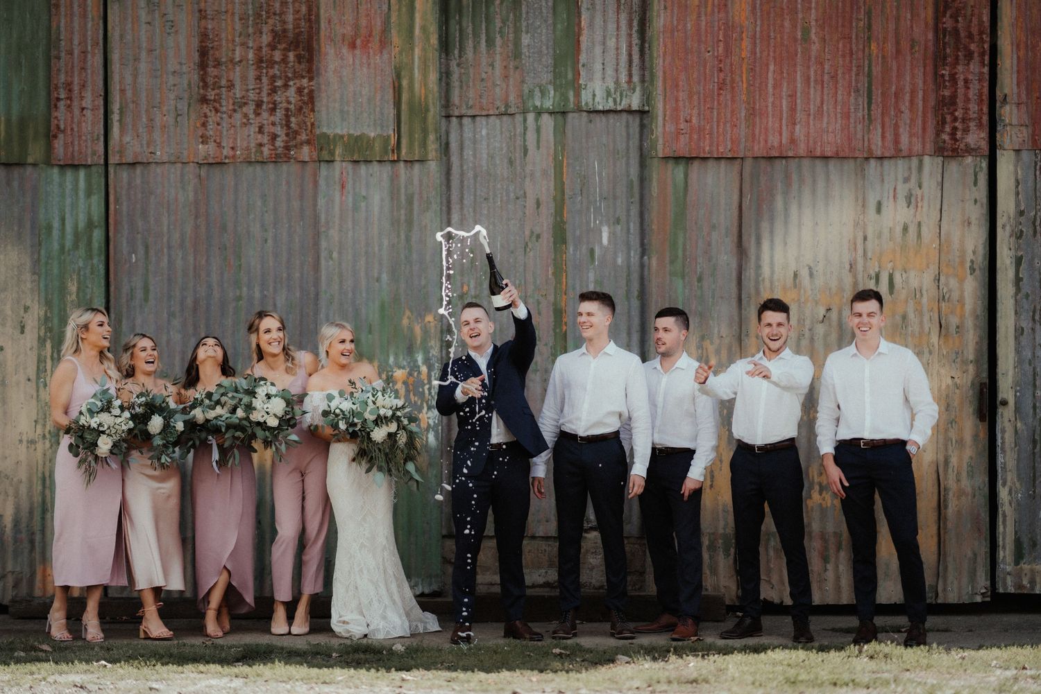 Bridal party poses in front of a rustic corrugated barn wall as the groom sprays champagne, with bridesmaids and groomsmen cheering.