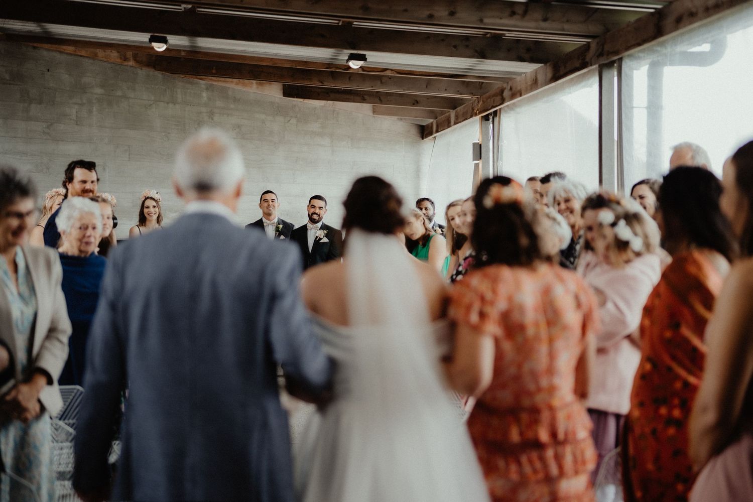 Wedding guests turn to watch as the bride walks down the aisle on her father's arm in a bright industrial-style venue.