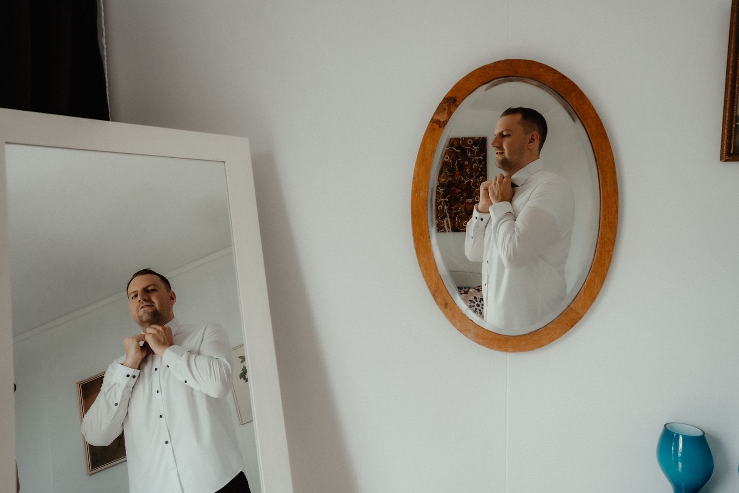 Groom adjusts his collar and bow tie while getting ready, seen in two mirrors on the morning of his wedding.