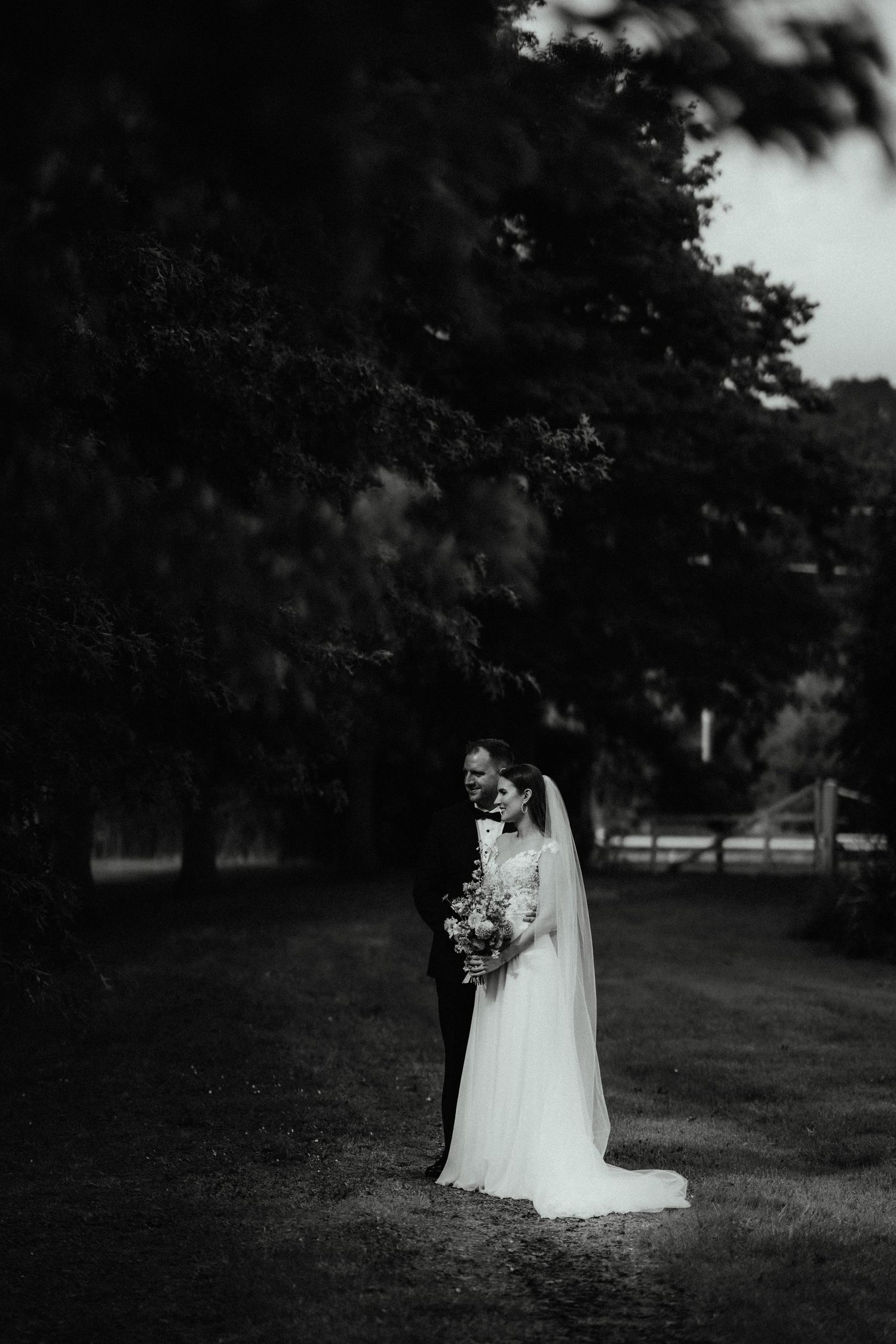 Black and white photo of a bride holding a bouquet and standing beside her groom under large trees in a grassy field.