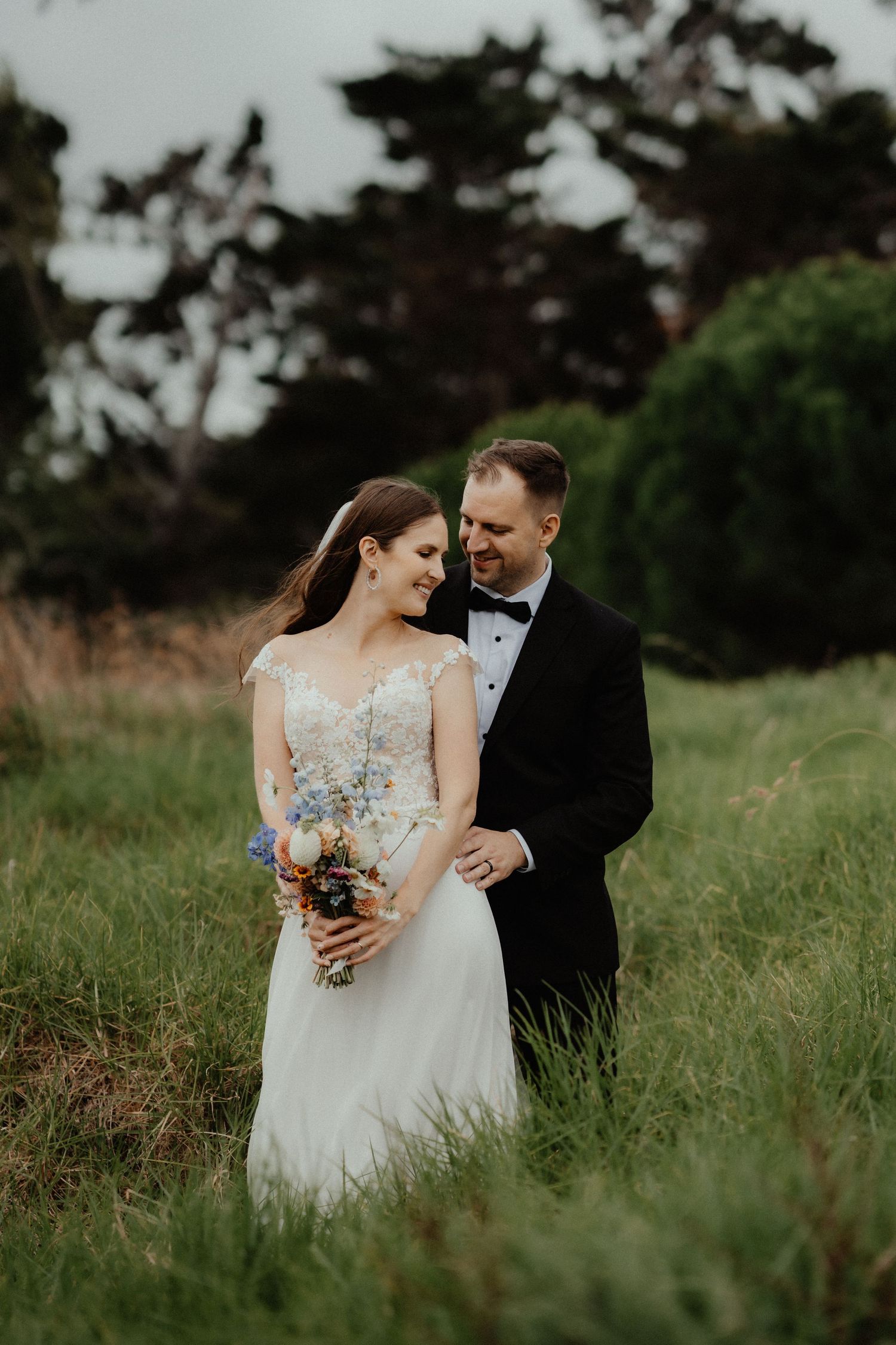 A smiling bride holding a colorful bouquet leans into her groom in a bow tie as they pose in a grassy meadow.