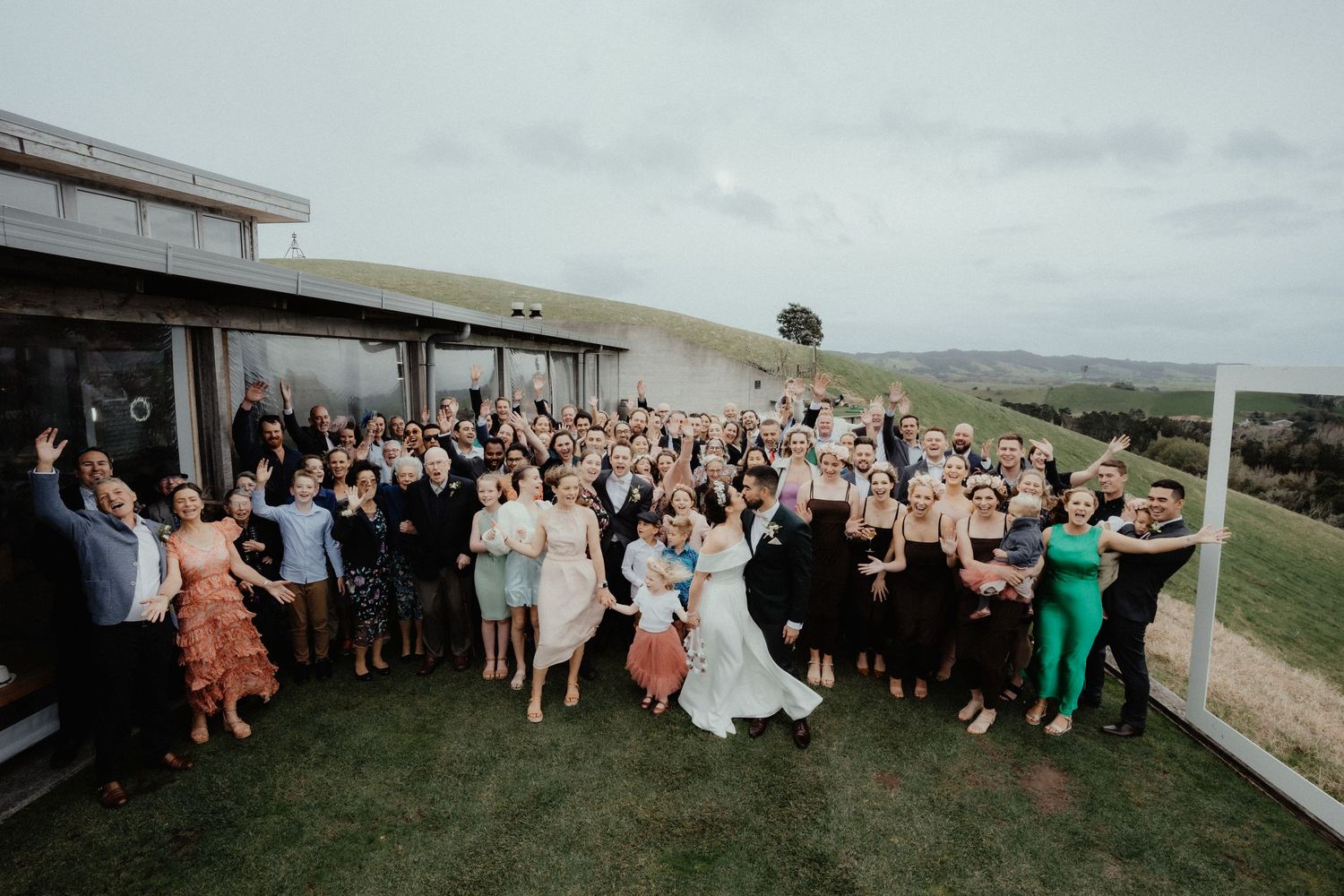 Large group of wedding guests gathers on the lawn outside a modern venue for a celebratory photo with the bride and groom.