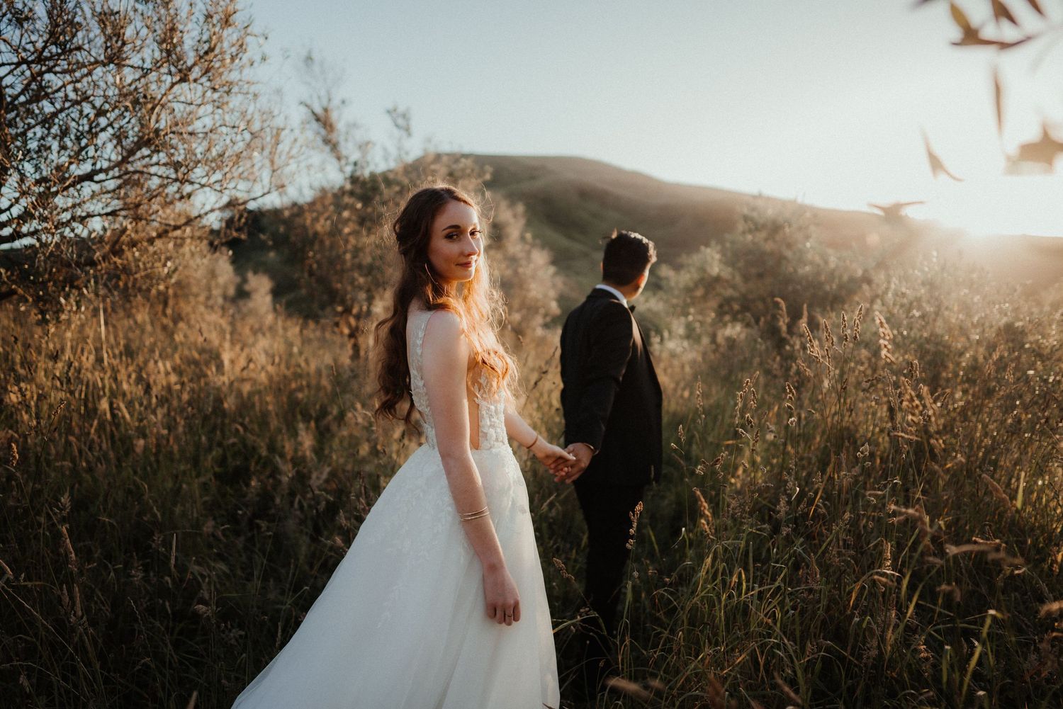 Bride in a white gown holds the groom's hand and looks back while walking through golden grass in warm sunset light.
