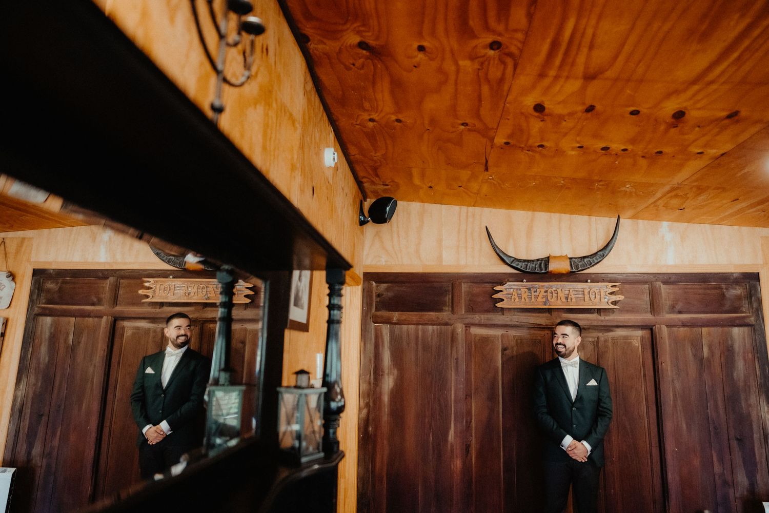 Two groomsmen in dark suits stand in a rustic wooden room with a longhorn skull mounted above the doors.