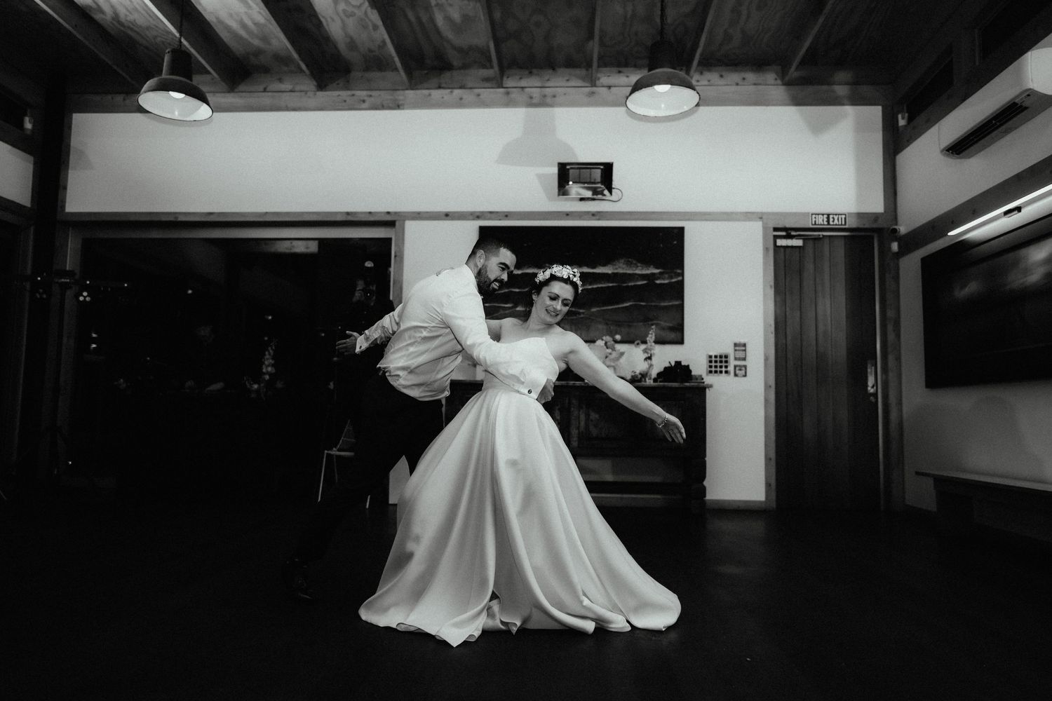 Black and white image of a groom dipping his bride during a first dance in a dim, rustic wedding venue.