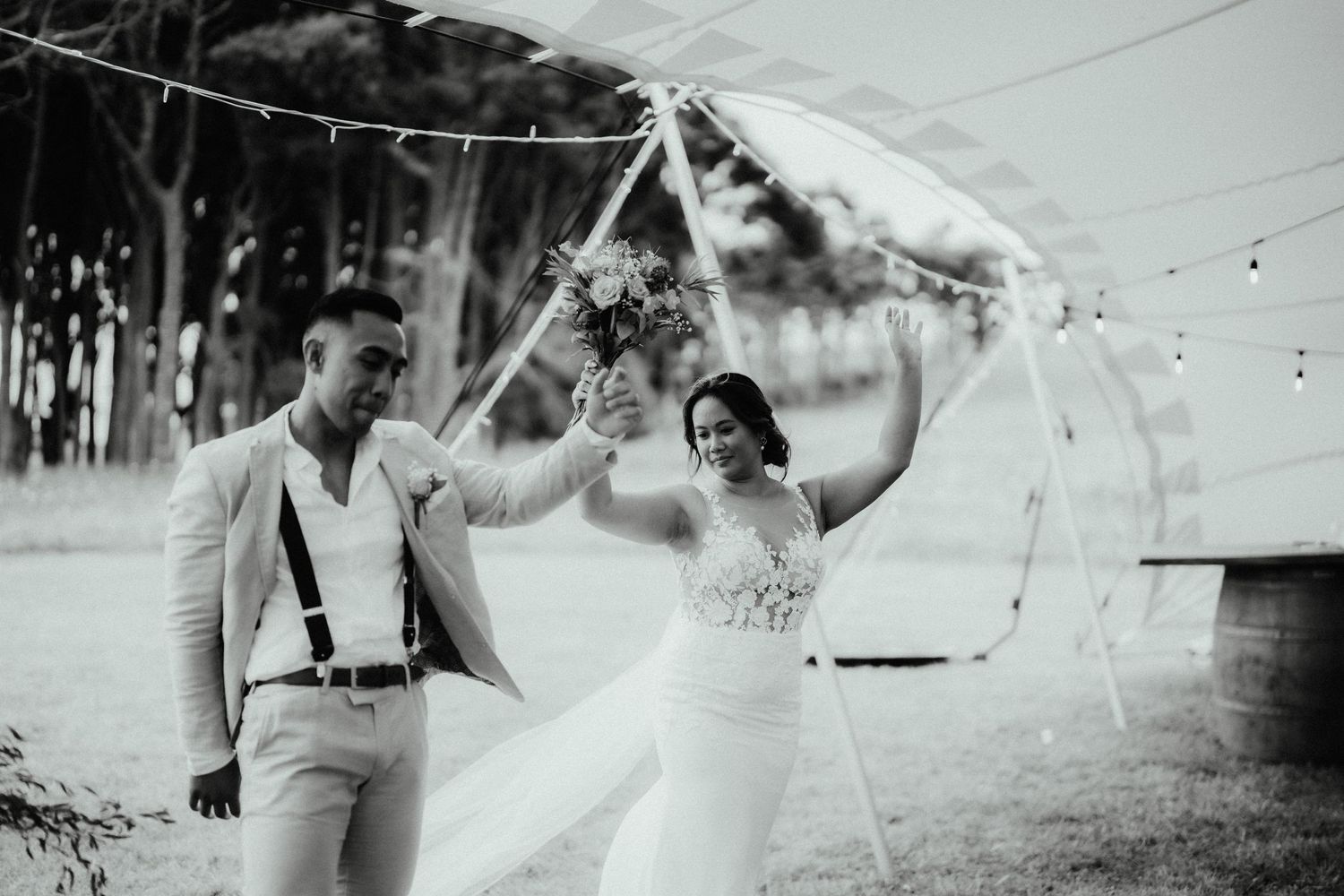 A smiling bride and groom walk hand in hand under a canopy tent with string lights, the bride holding a bouquet aloft.
