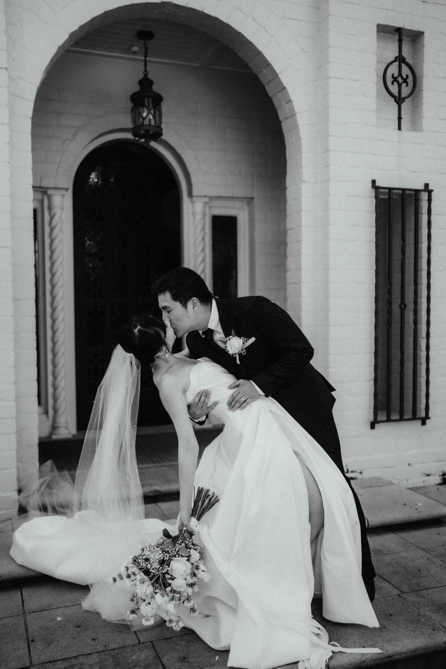 Black and white photo of a groom dipping and kissing his bride in a veil and satin gown outside an arched doorway.