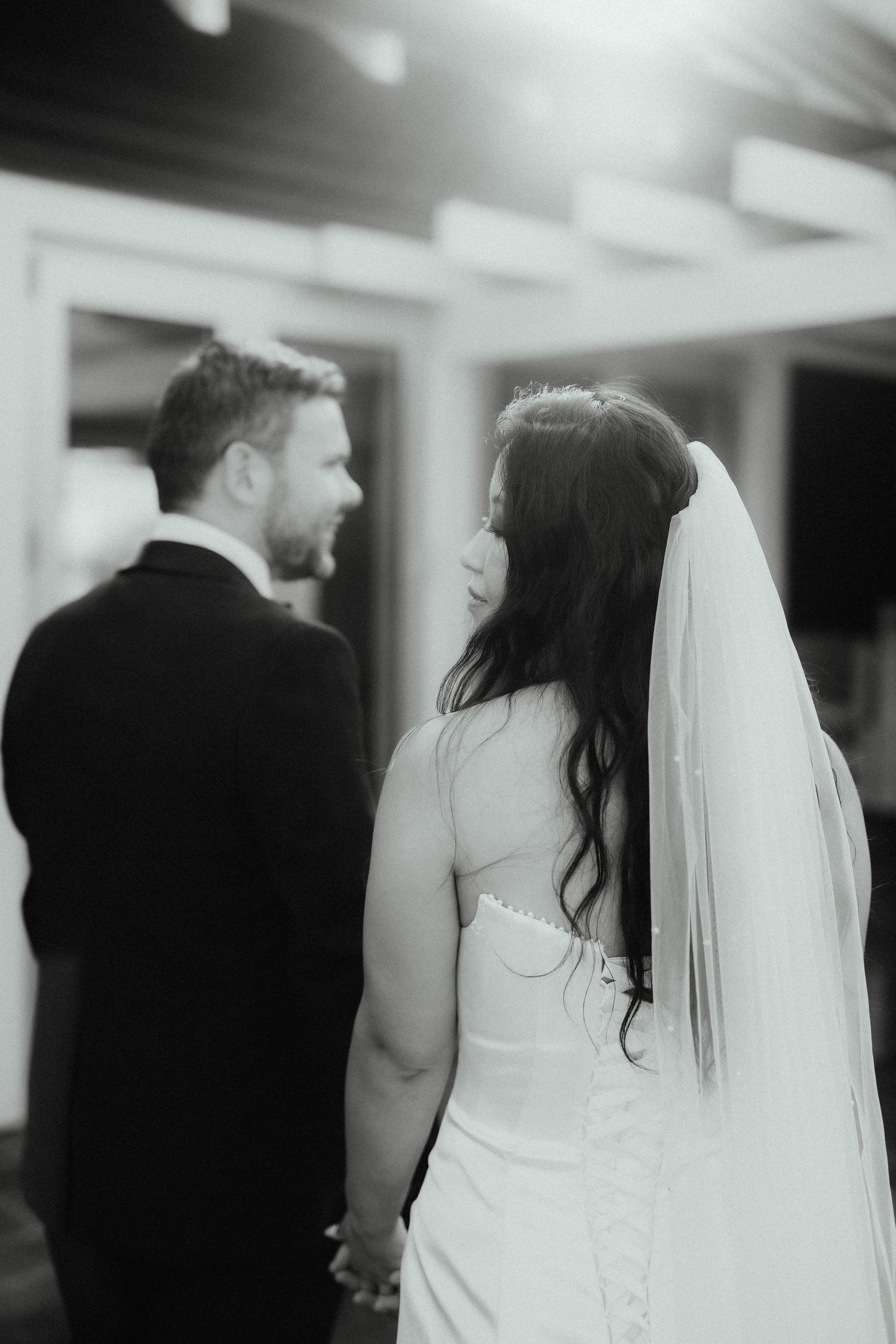 Black and white photo of a bride in a long veil holding hands with her groom as they look back at each other.