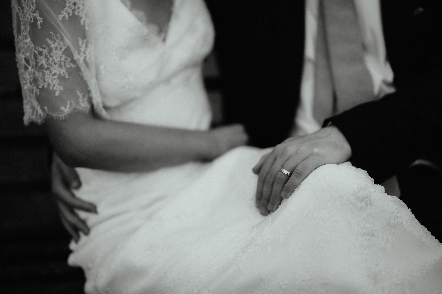 Black and white close-up of a bride in a lace wedding dress with a groom's hand wearing a ring resting gently on her lap.