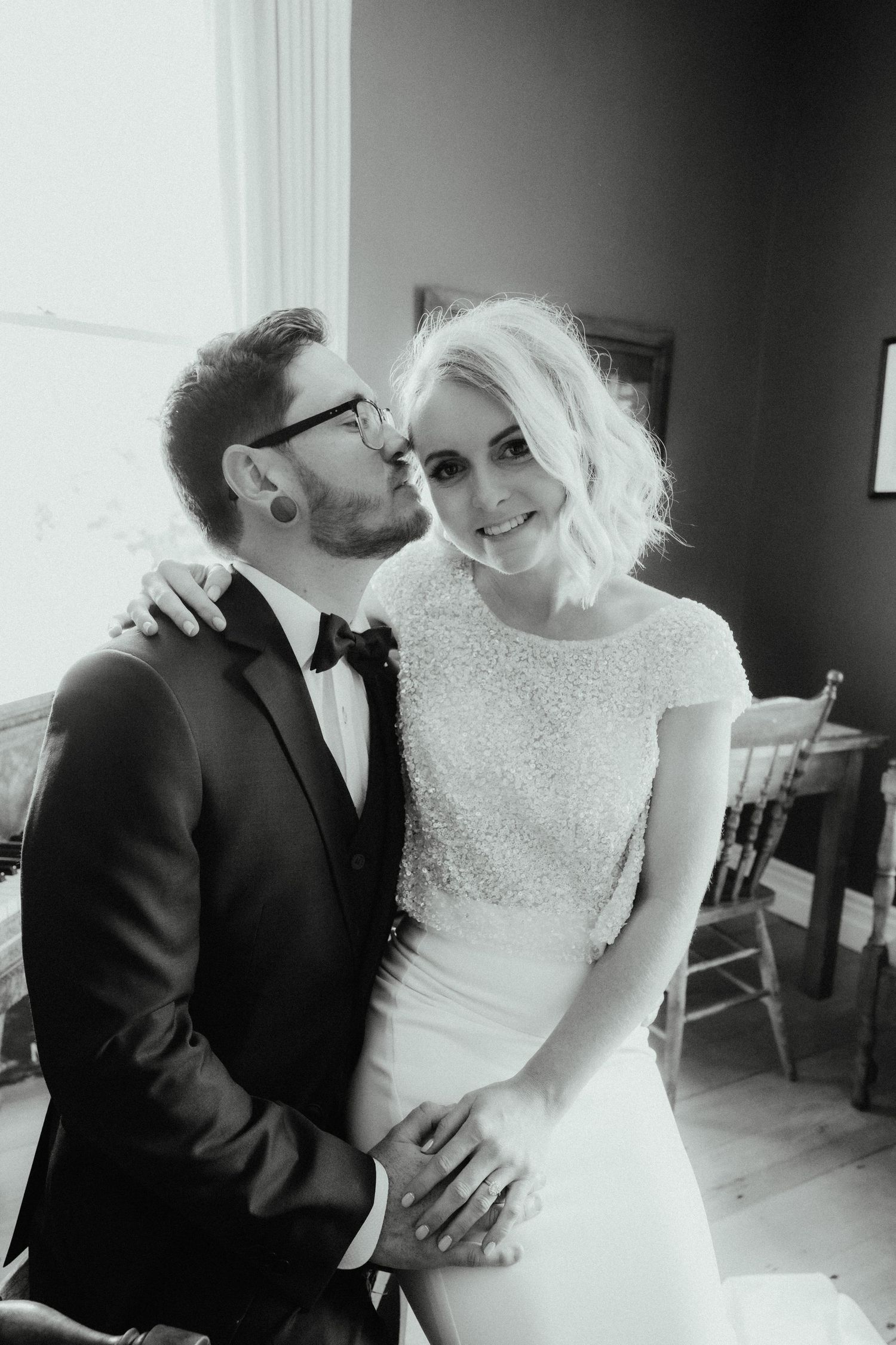 Black and white photo of a groom in glasses and bow tie kissing the cheek of his smiling bride seated beside him.