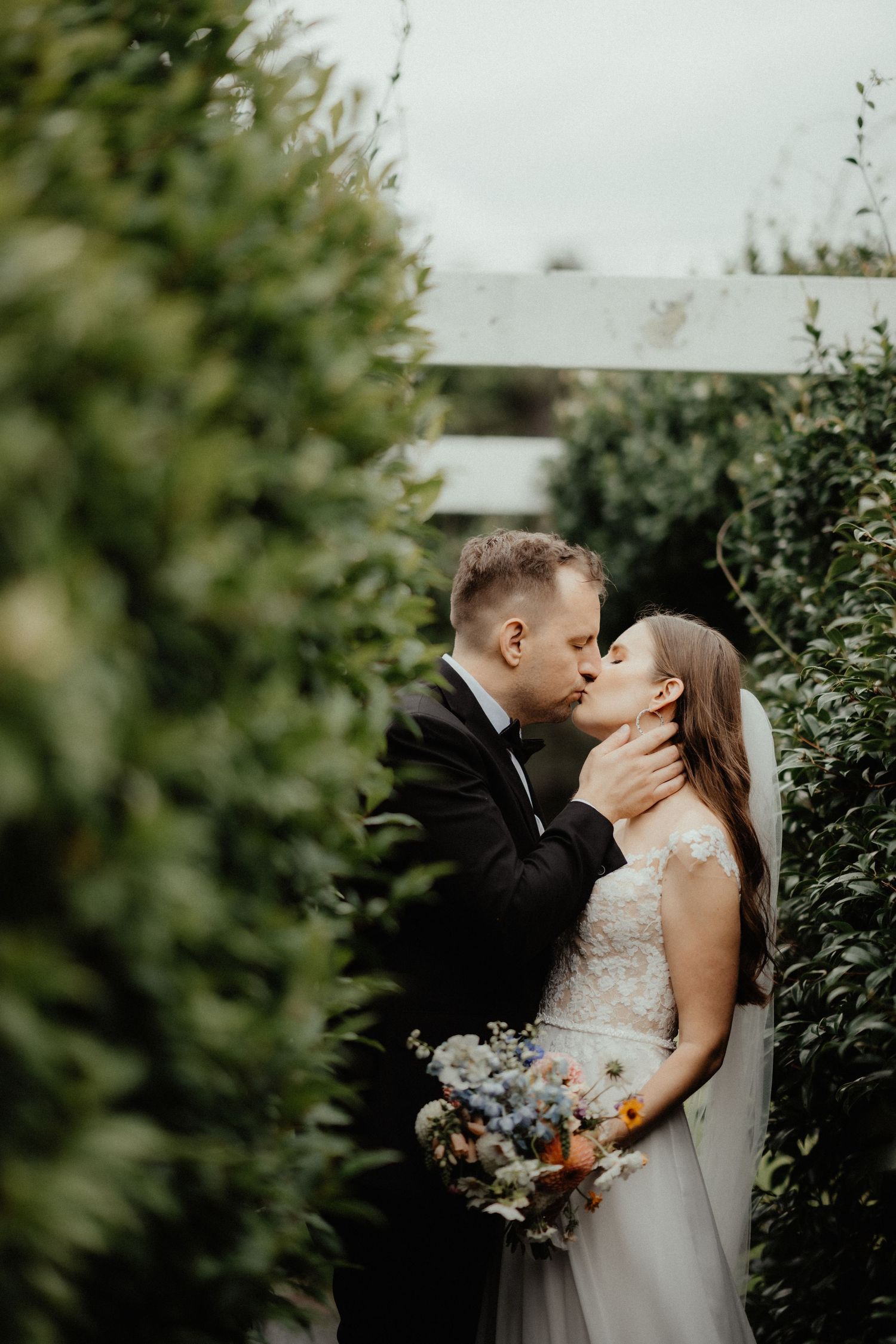 Groom in tuxedo kisses the bride holding a blue and white bouquet beside a tall green hedge and white pergola.