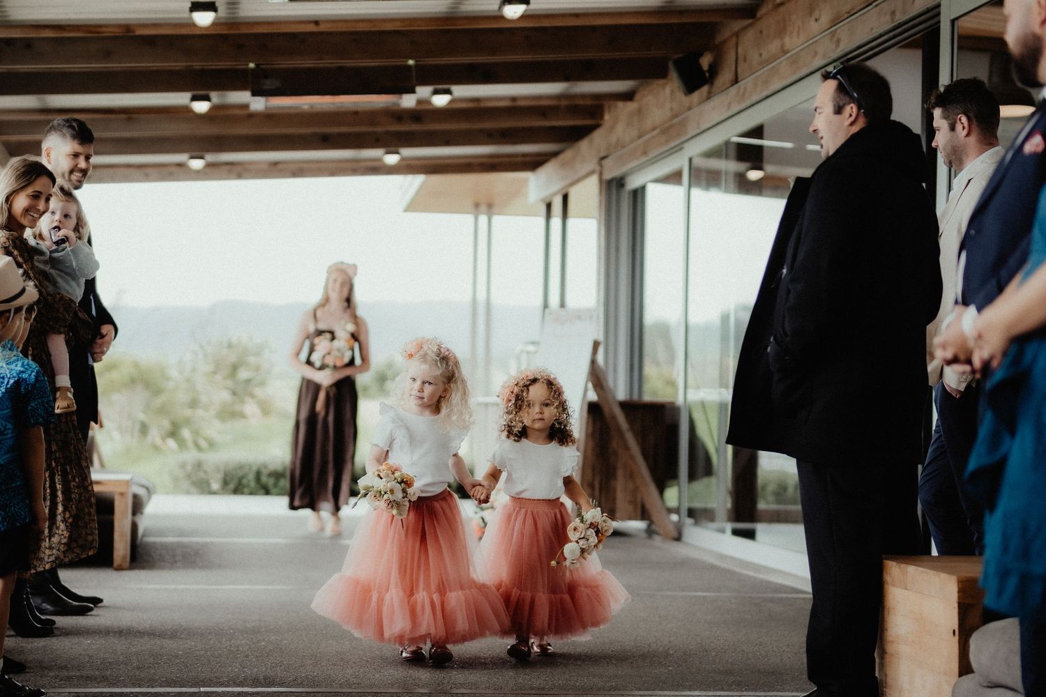 Two flower girls in pink tulle skirts walk down the aisle holding bouquets as wedding guests look on in an open-air venue.