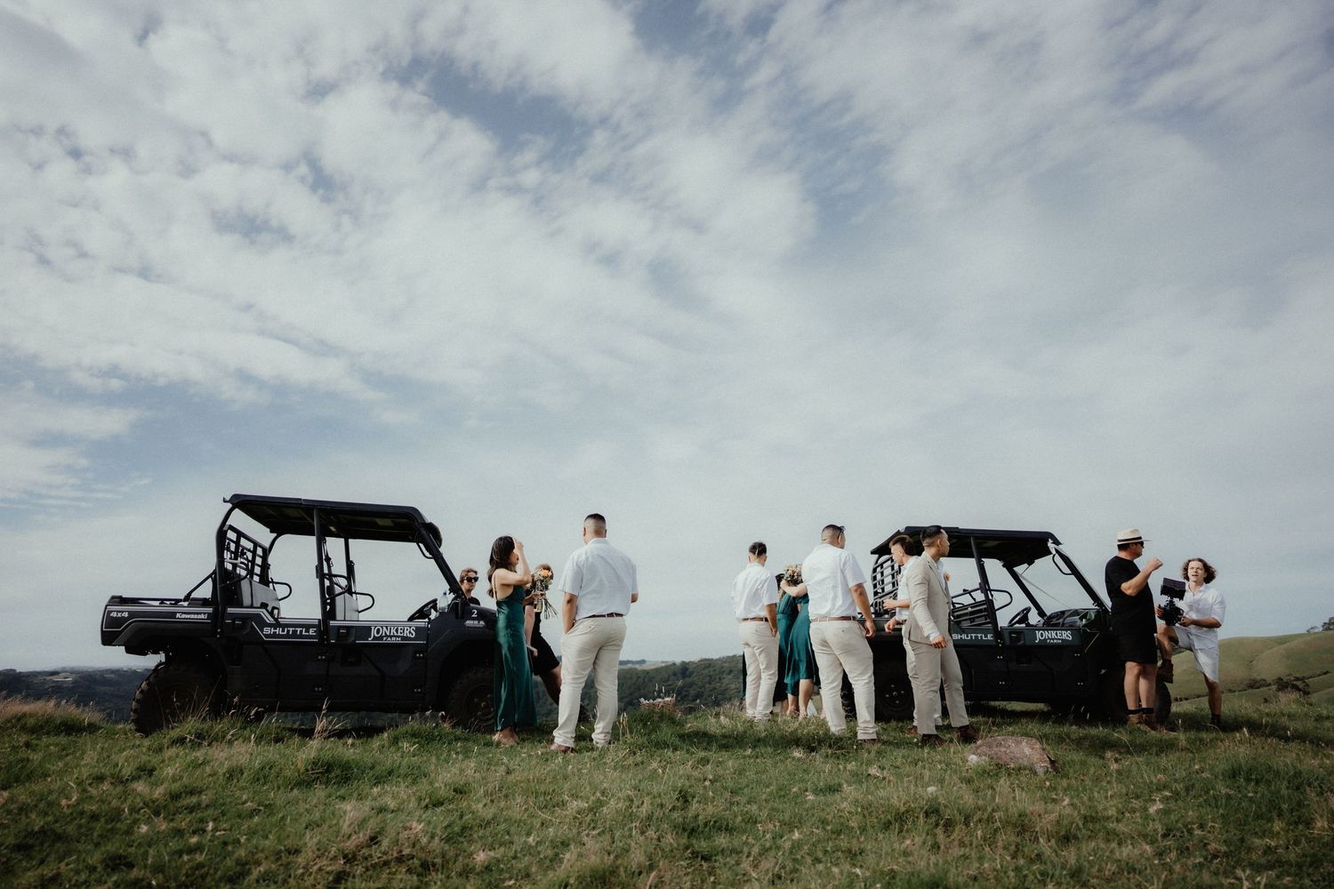Wedding guests in light attire gather beside two black utility vehicles on a grassy hilltop under a cloudy sky.