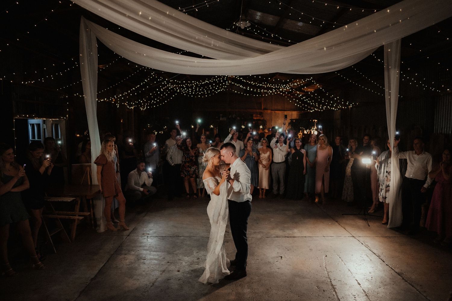 Newlyweds share their first dance under draped fabric and string lights while guests hold up lights in a barn venue.