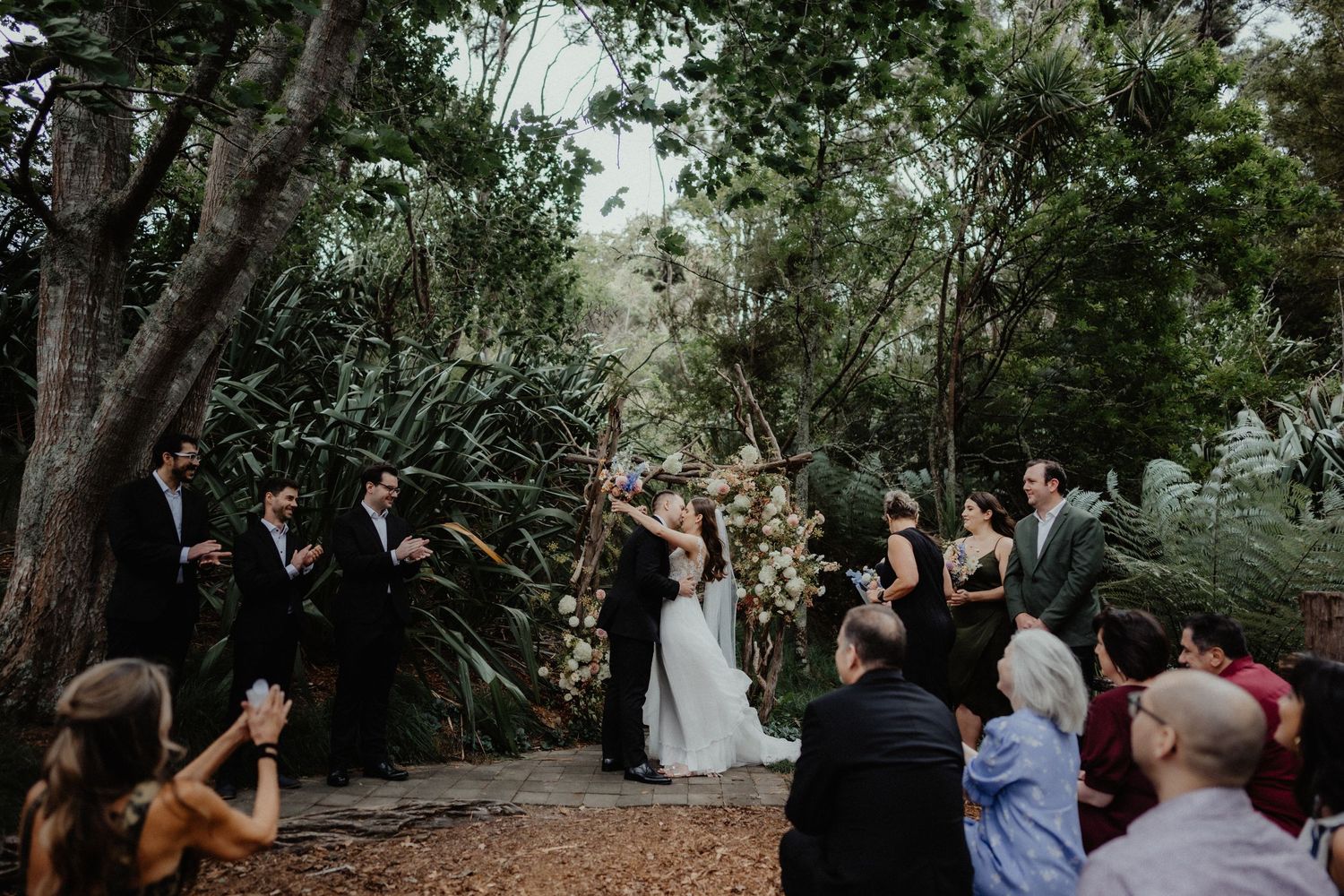 Wedding guests watch as the bride and groom share a moment during an outdoor ceremony surrounded by lush native bush.
