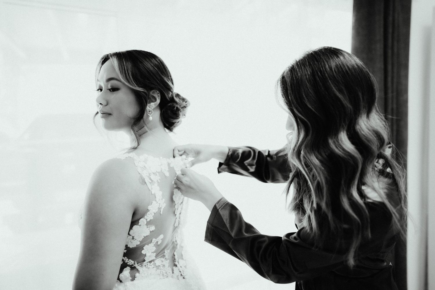 Black and white photo of a bridesmaid fastening the lace back of the bride's wedding dress by a bright window.