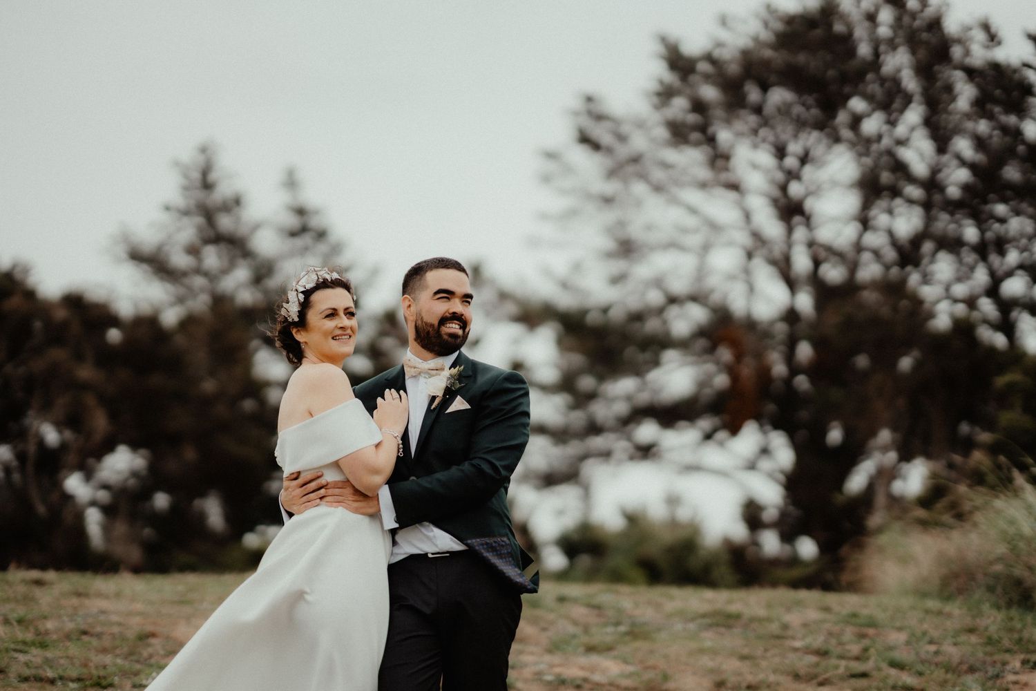 Bride and groom embrace and smile outdoors on a grassy hill with tall trees behind them on a cloudy day.