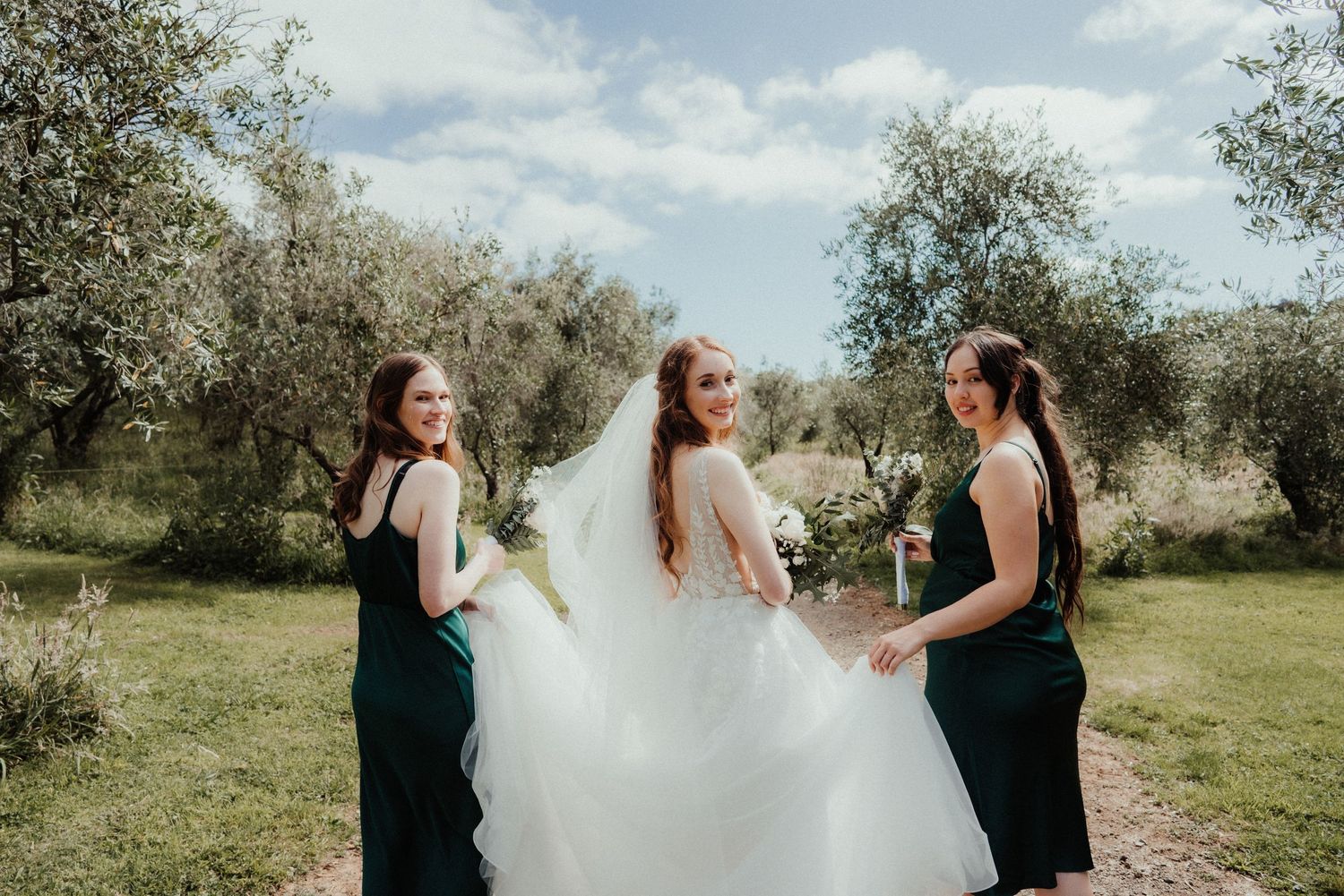 Two bridesmaids in dark green dresses hold the bride's flowing white gown while walking through a sunny olive grove.