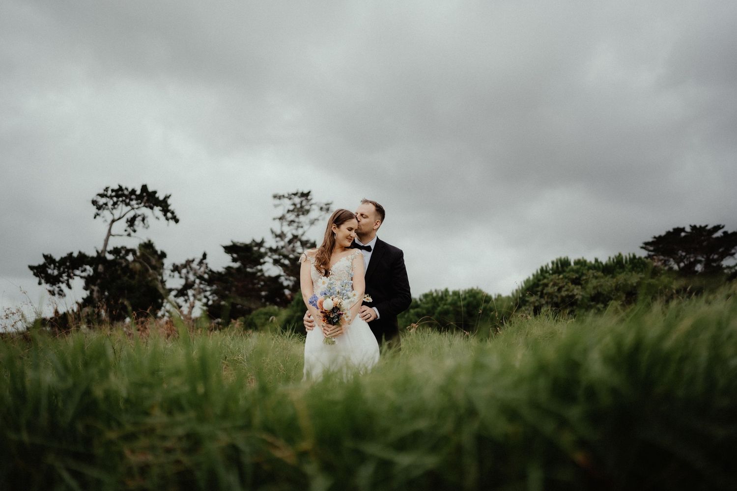 Bride and groom embrace in a grassy field beneath a moody, overcast sky at their outdoor wedding.