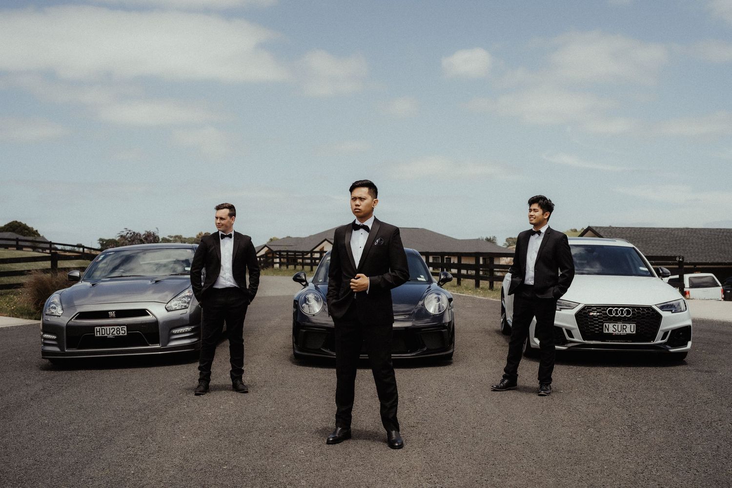 Groom in a black tuxedo stands in front of two groomsmen and three luxury cars on a driveway under a cloudy sky.