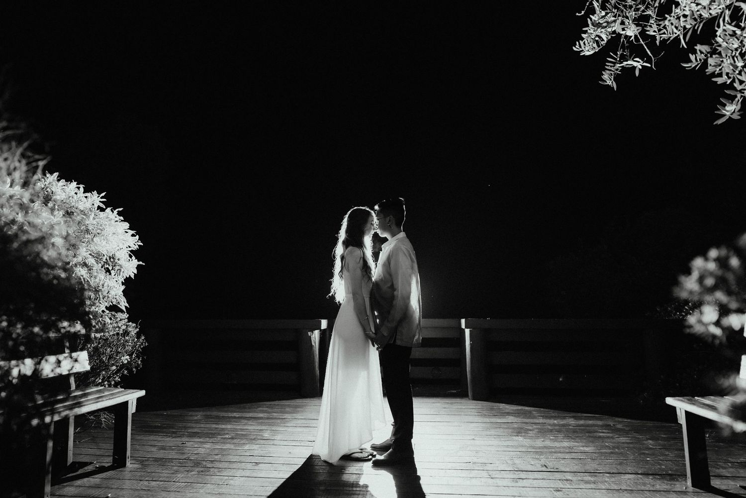 Silhouetted bride and groom share an intimate moment on a wooden deck at night in a black and white wedding photo.
