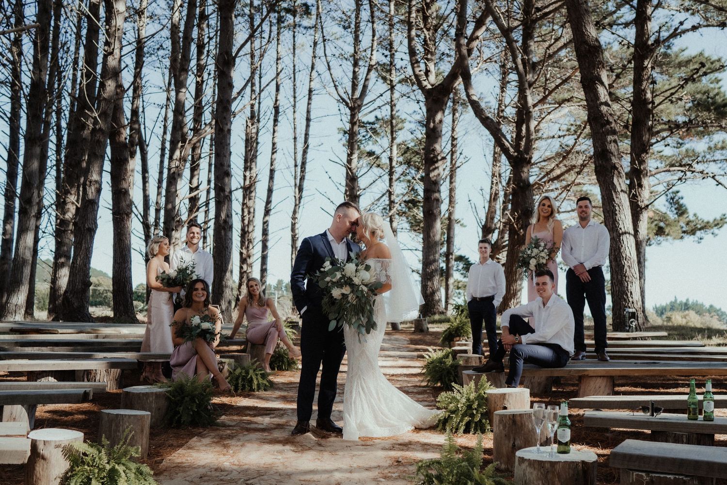 Newlyweds hold a bouquet and kiss on a forest aisle lined with wooden benches as the wedding party smiles beside them.