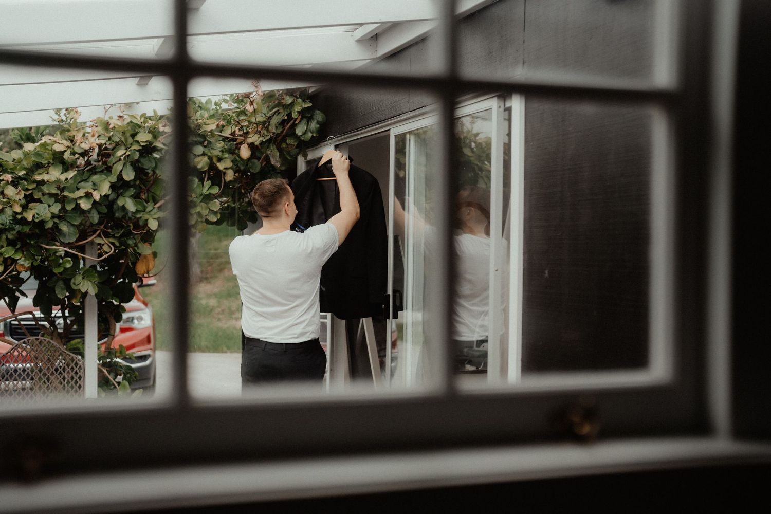 Groom in a white shirt hangs his suit jacket near a window, viewed through a window pane during wedding morning preparations.