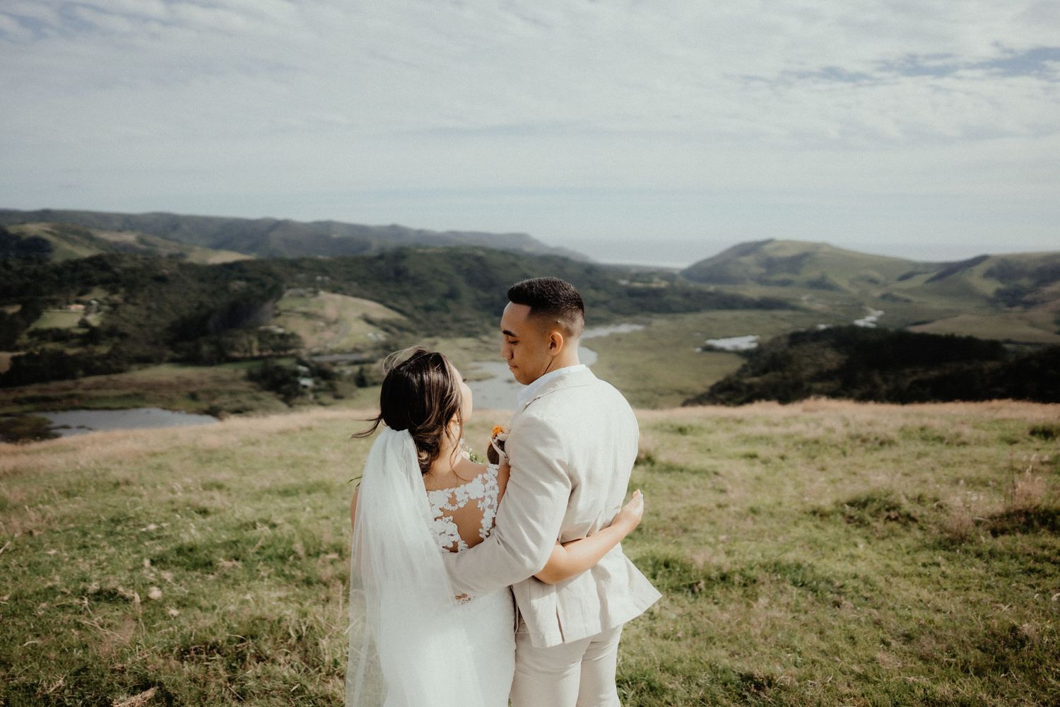Bride in a long veil and groom in a cream suit embrace on a grassy hilltop overlooking valleys and distant hills.